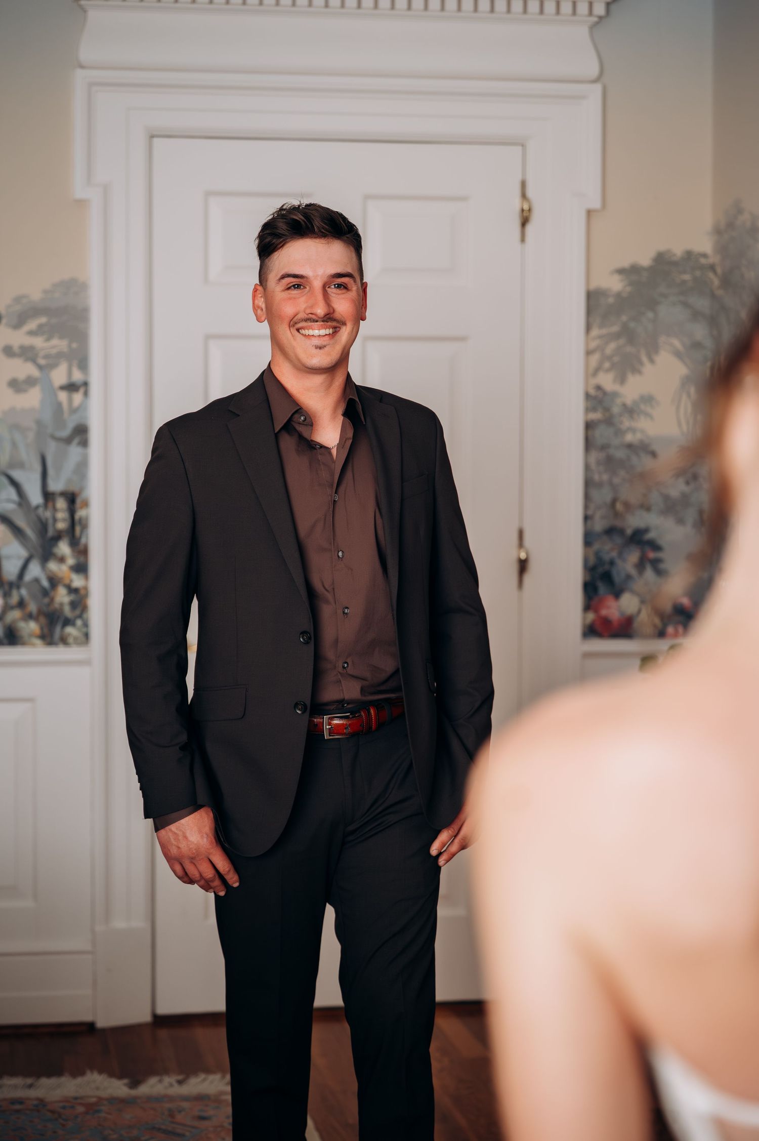 Smiling groom in black suit and dark shirt stands confidently in elegant room with floral wallpaper.