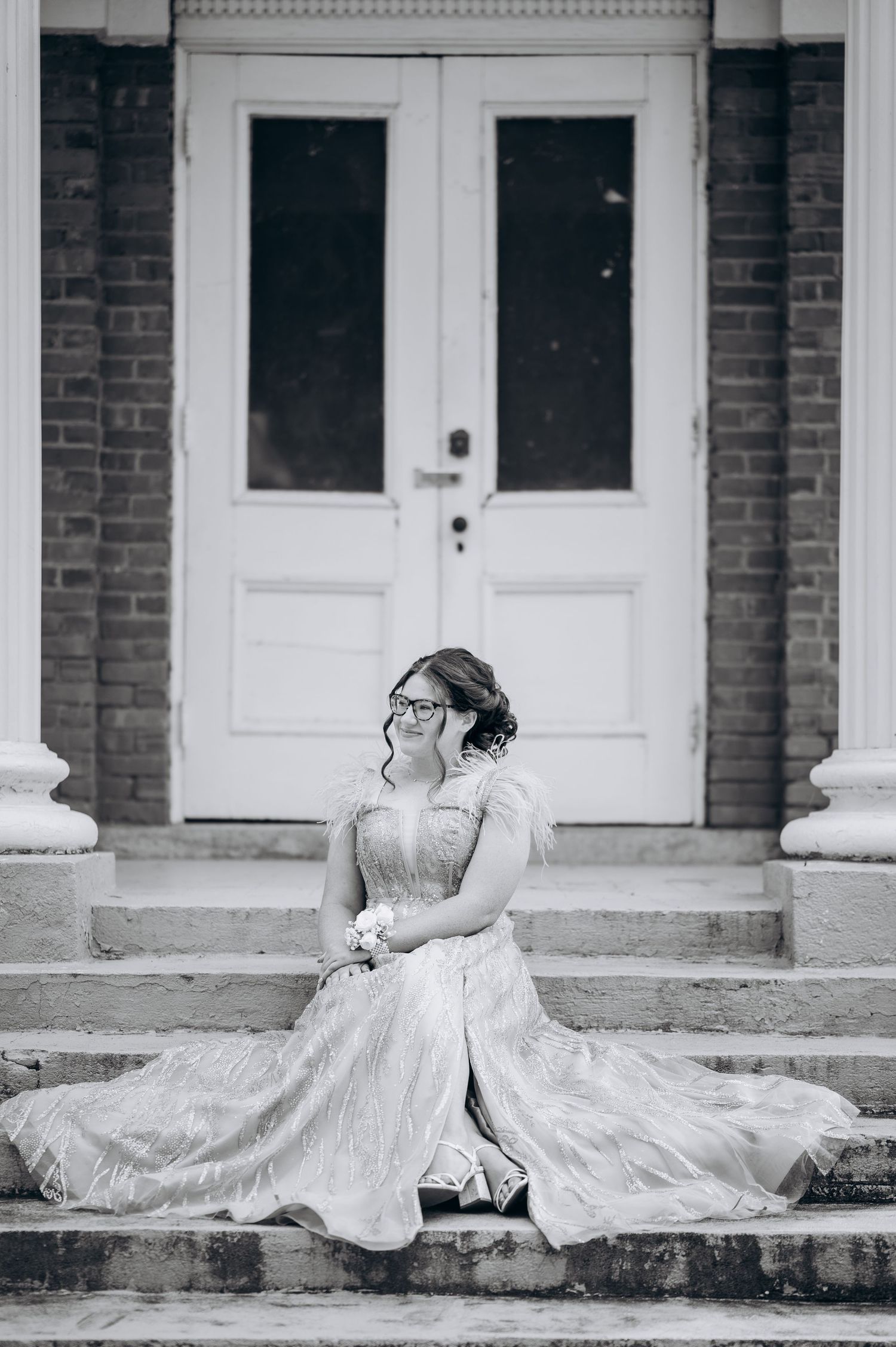 Bride in white gown and glasses sits on stone steps before white double doors in elegant black and white portrait.