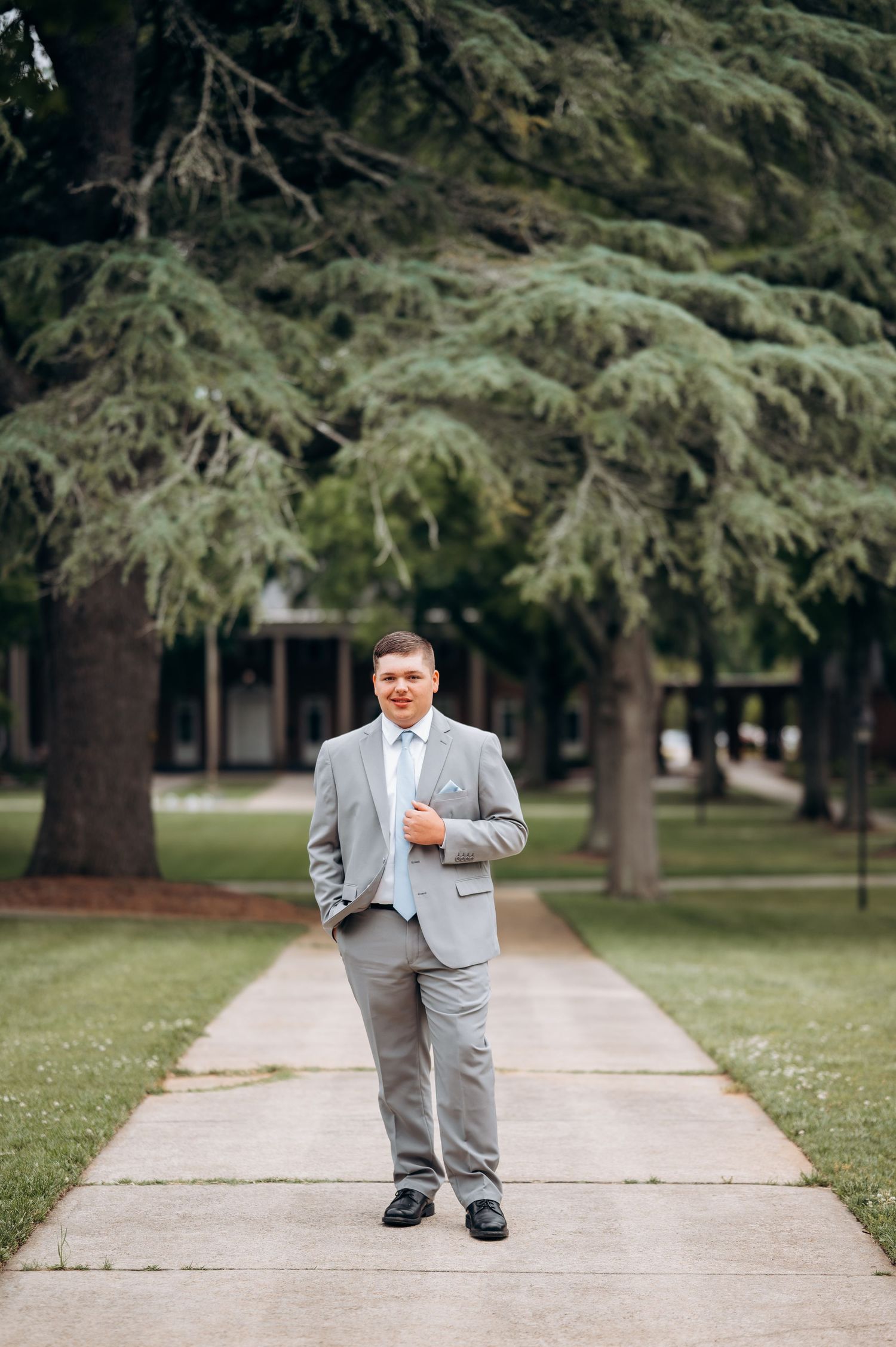 A groom in a light gray suit stands confidently on a tree-lined pathway, smiling outdoors.
