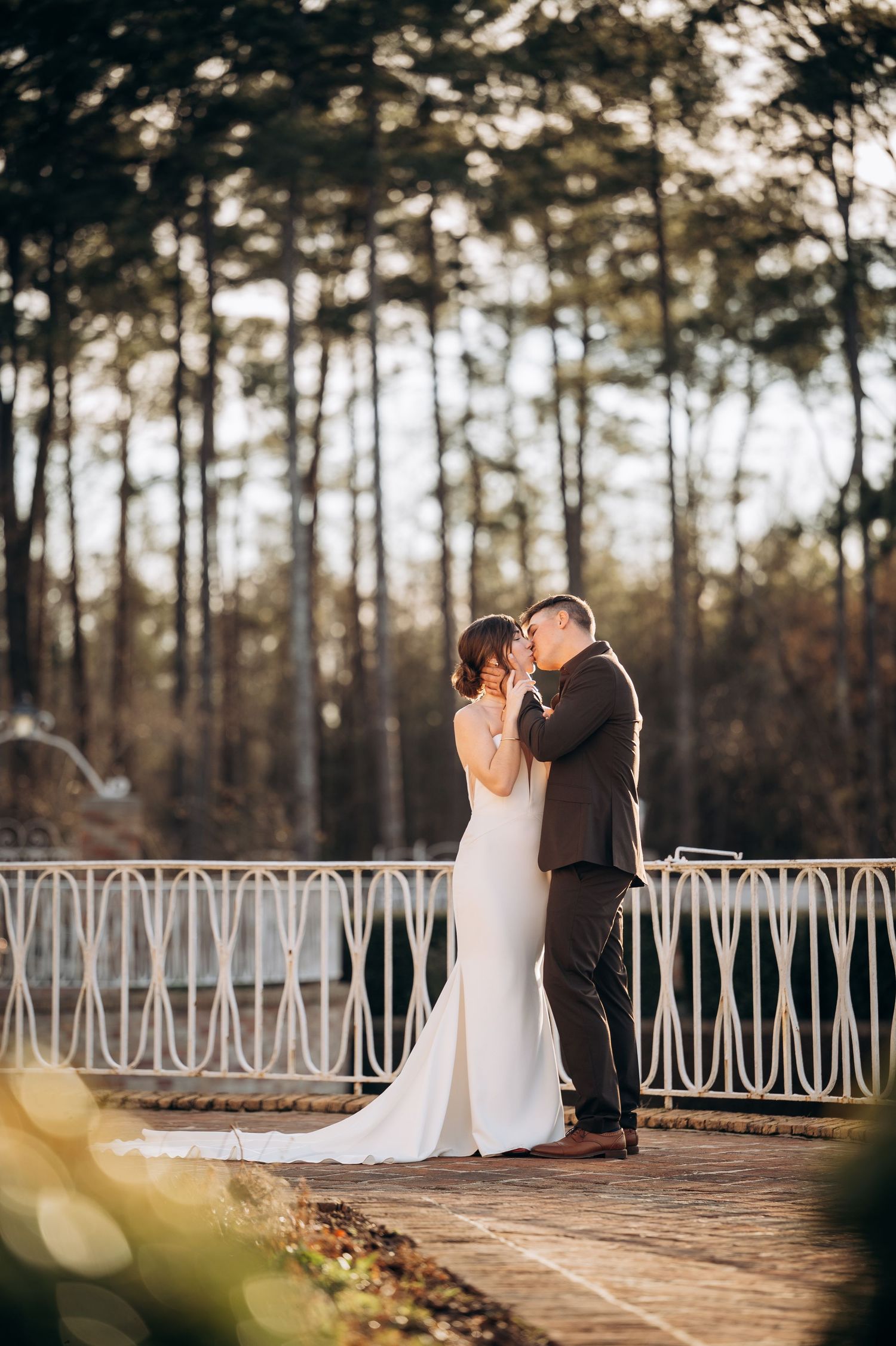 Couple in wedding attire share a romantic moment on a pathway with decorative white railings at sunset.