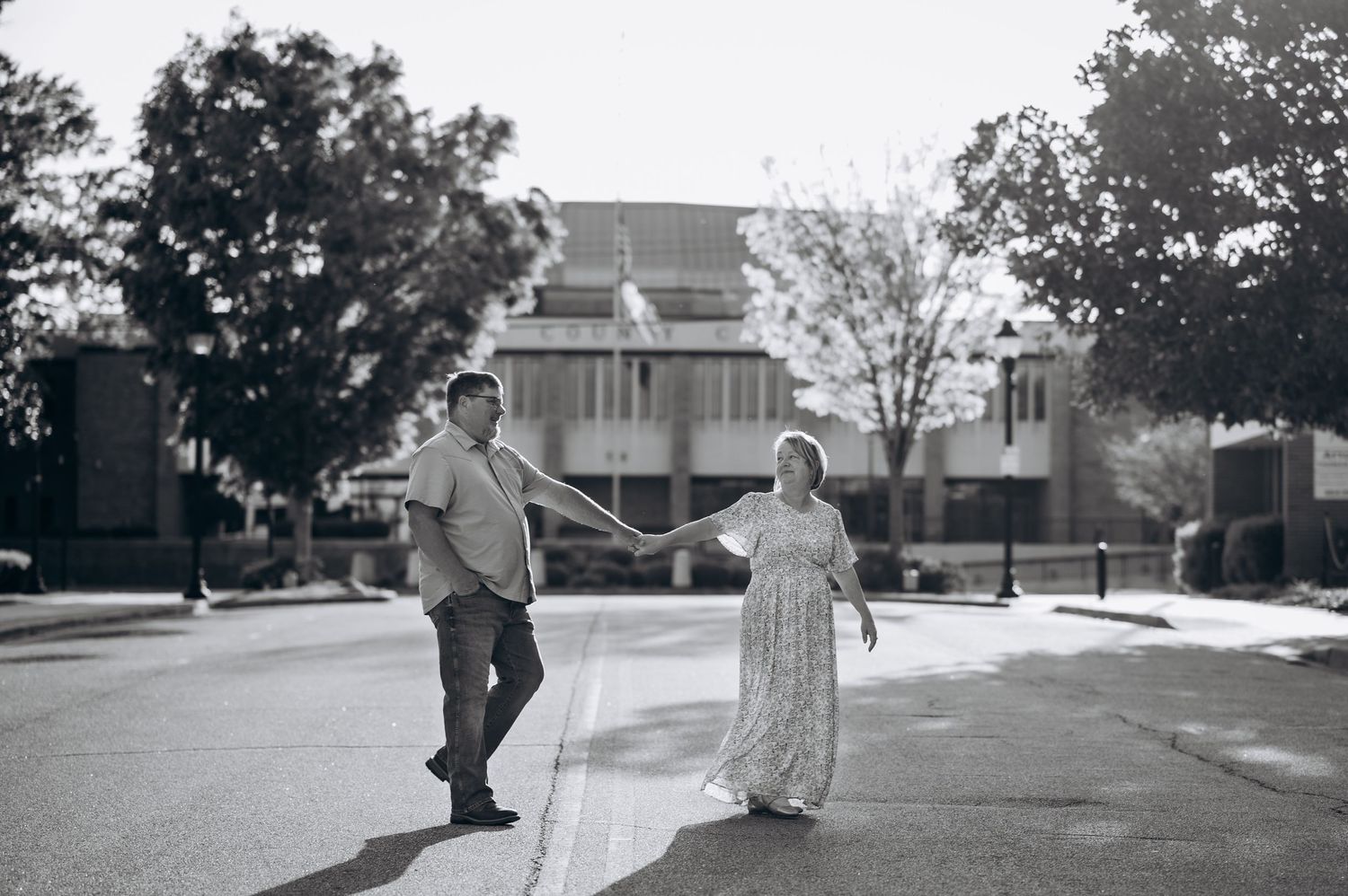 Black and white photo of a couple holding hands while crossing a sunny street, with trees and buildings in the background.