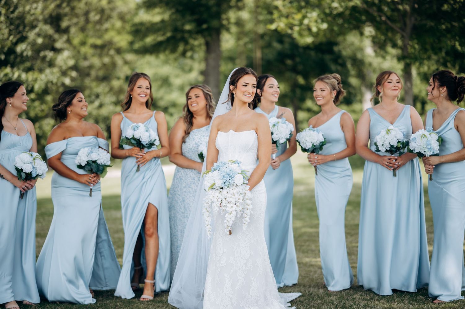 Bride in white lace gown with bridesmaids in light blue dresses holding white bouquets outdoors.
