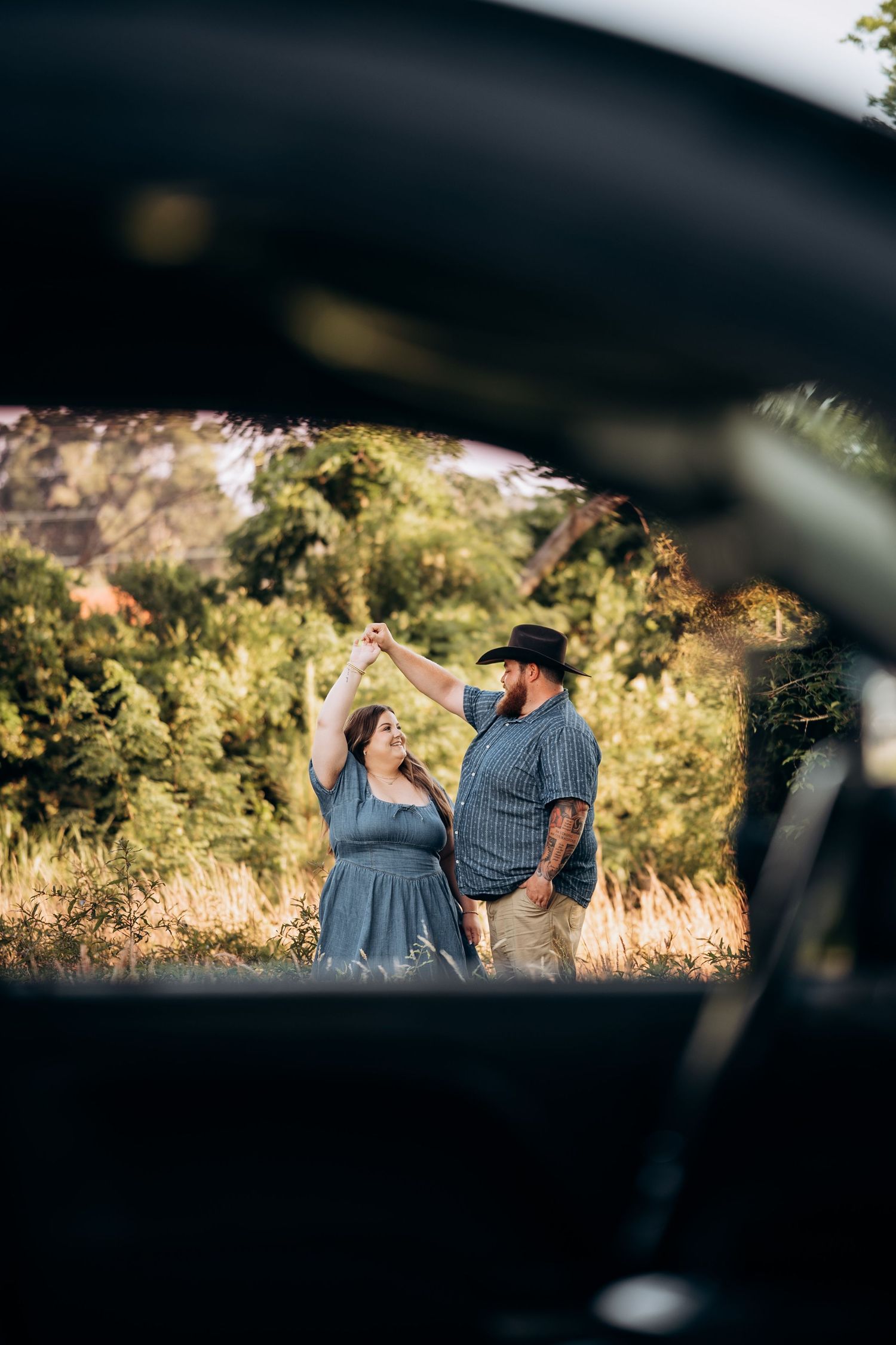 Couple dancing in a sunlit green field, photographed through a truck window frame.