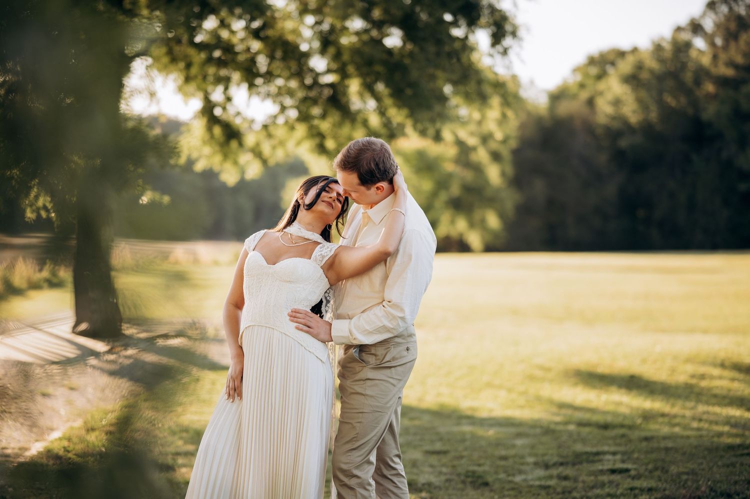 Bride and groom share a romantic moment in a sunlit park, bride in white gown, groom in tan suit.