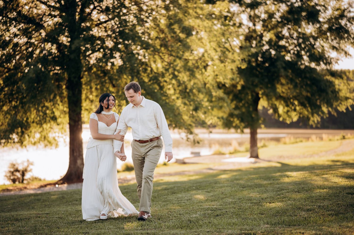 Bride and groom walking hand in hand on lush green lawn near lake at golden hour wedding celebration.