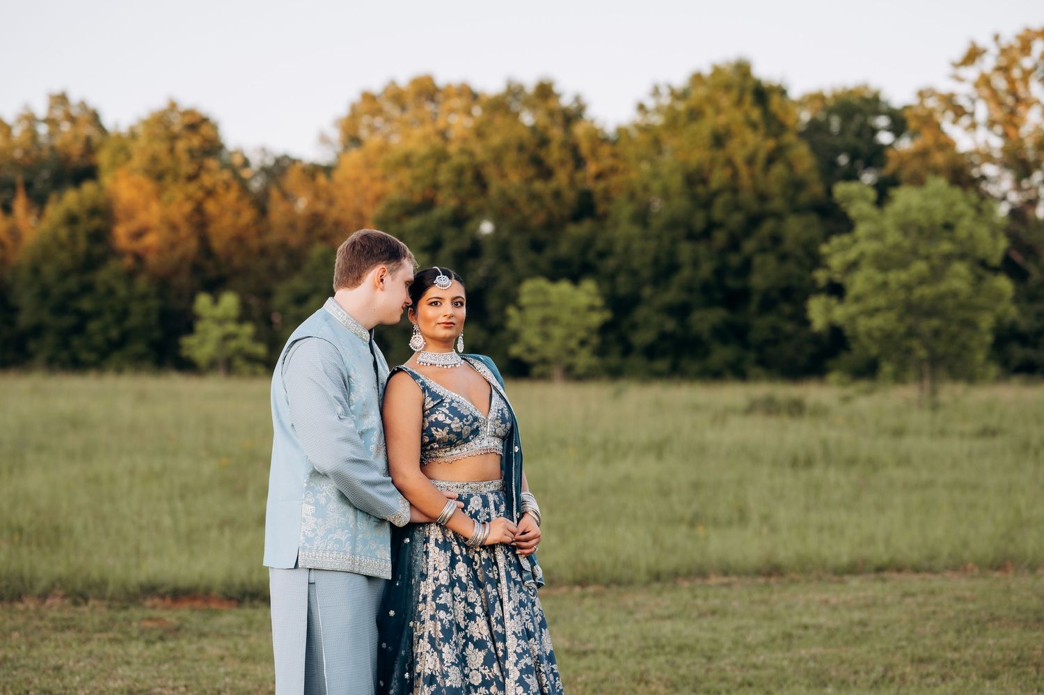 Couple in elegant attire stands in green field at sunset, woman in blue floral lehenga, man in light gray suit.