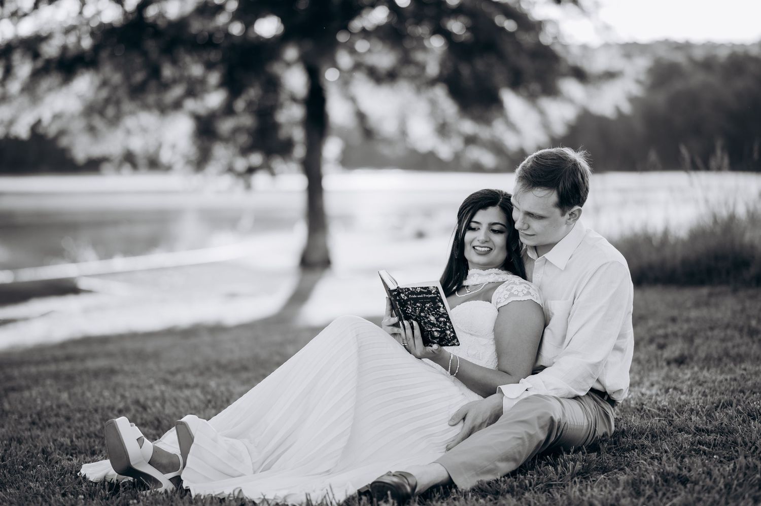 Black and white photo of a couple sitting on grass by a lake, reading a book together in a romantic outdoor setting.