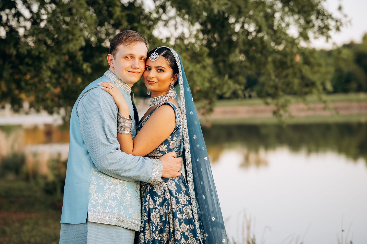 Couple in blue Indian wedding attire embracing outdoors near a lake, blending Western and South Asian styles.