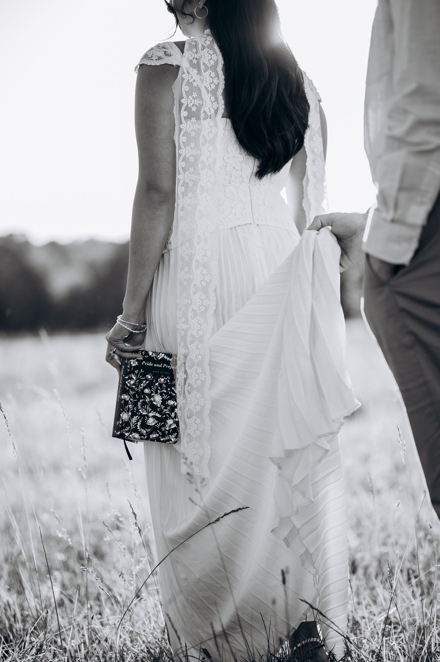 Black and white photo of a bride in lace wedding dress holding a clutch, standing in a field with groom.