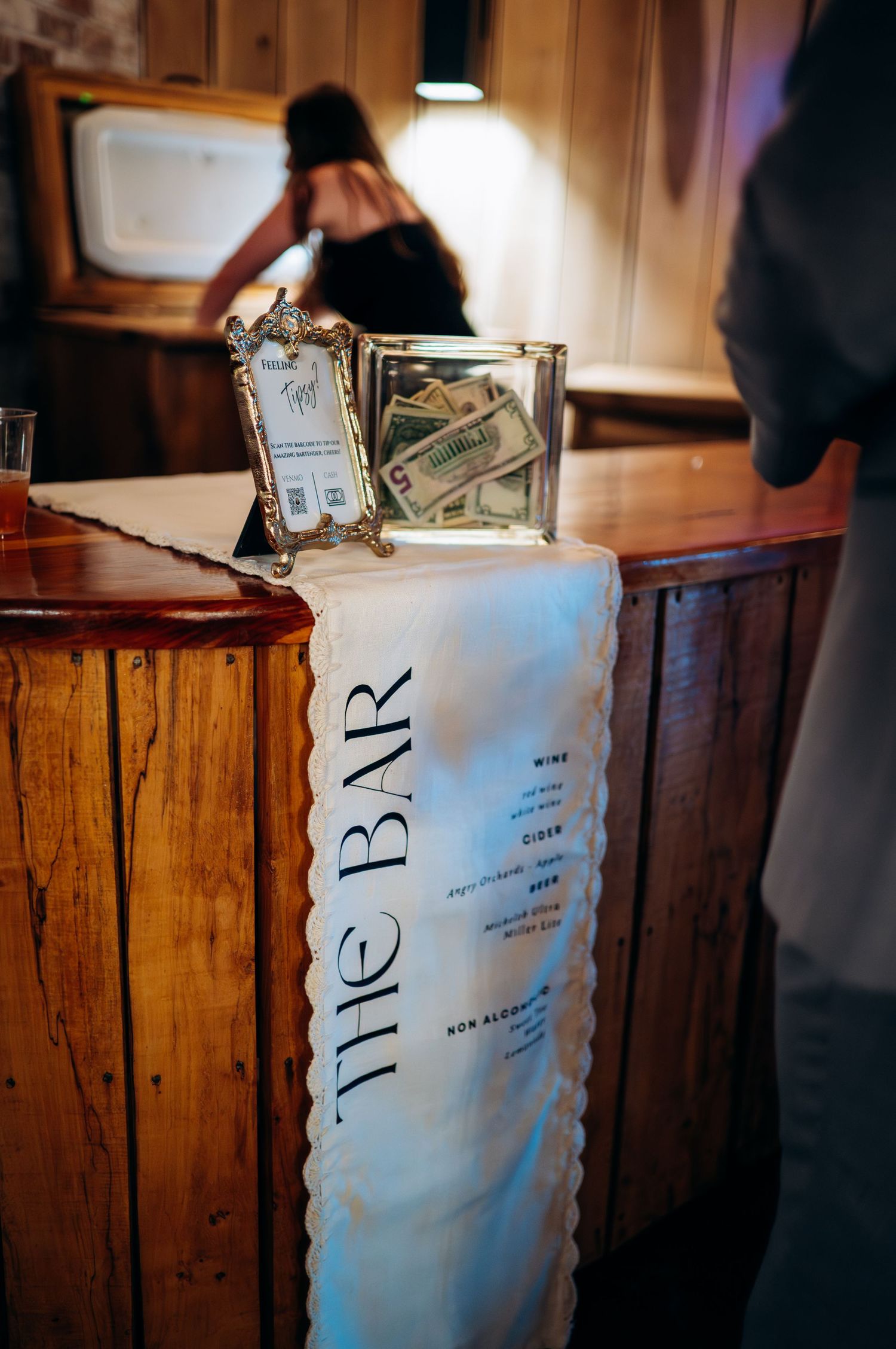 Elegant wedding bar setup with tip jar and decorative white linen bar sign.