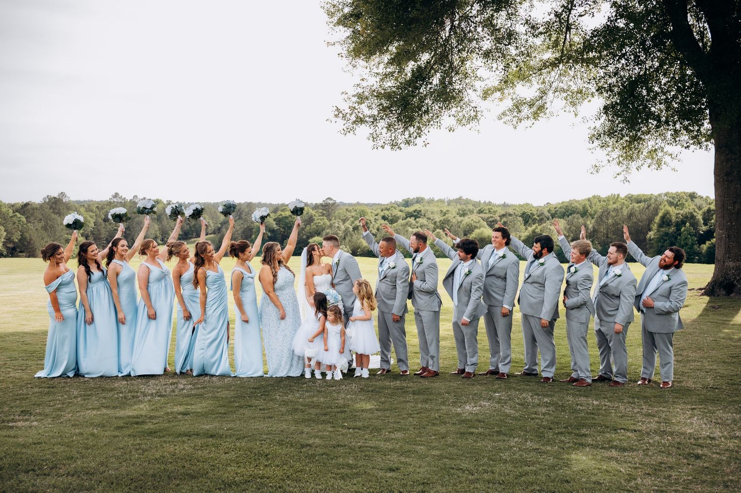 Large wedding party in light blue dresses and gray suits celebrate outdoors under tree on lawn.