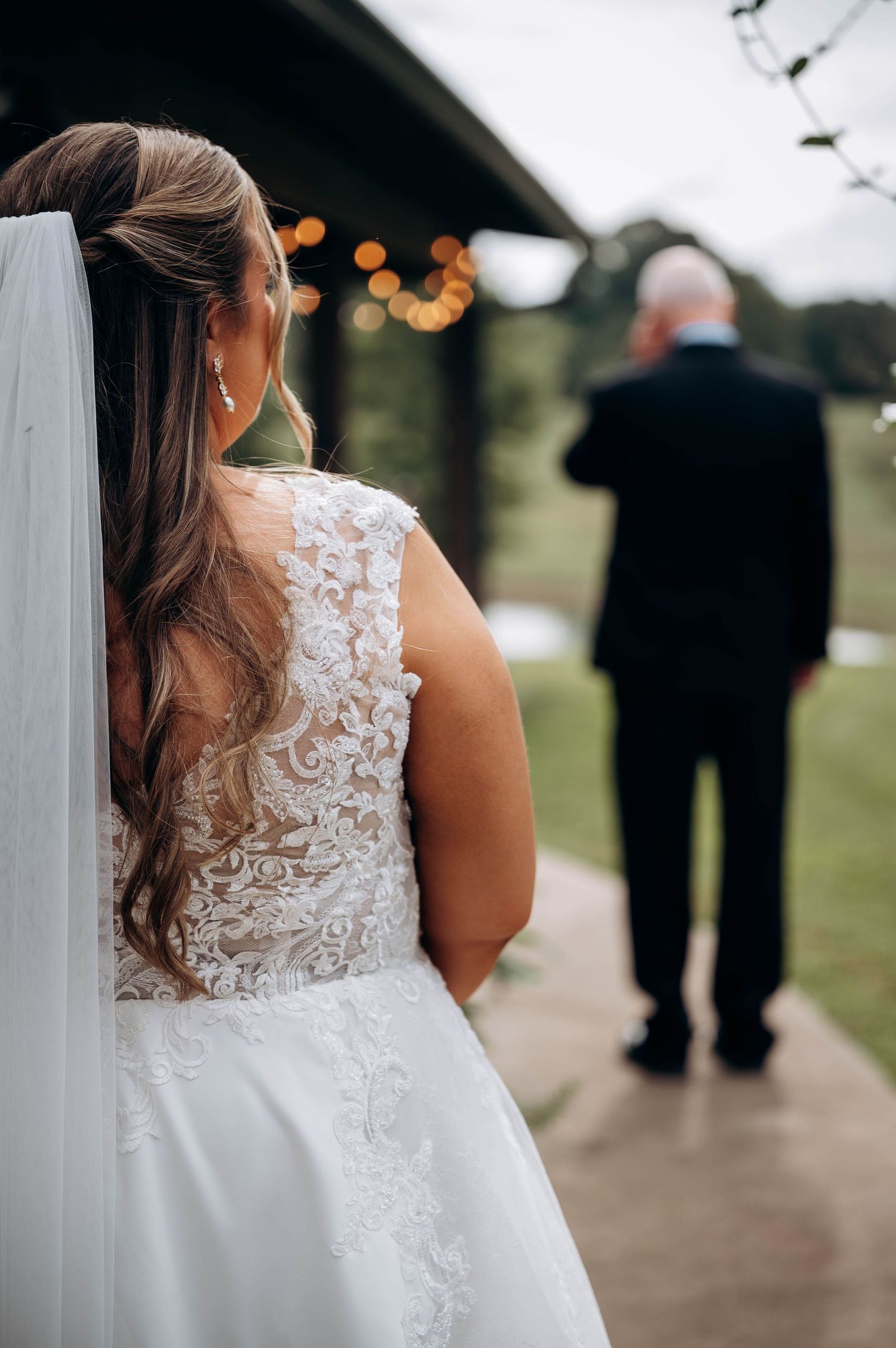 Bride wearing detailed lace back wedding gown stands on covered pathway.