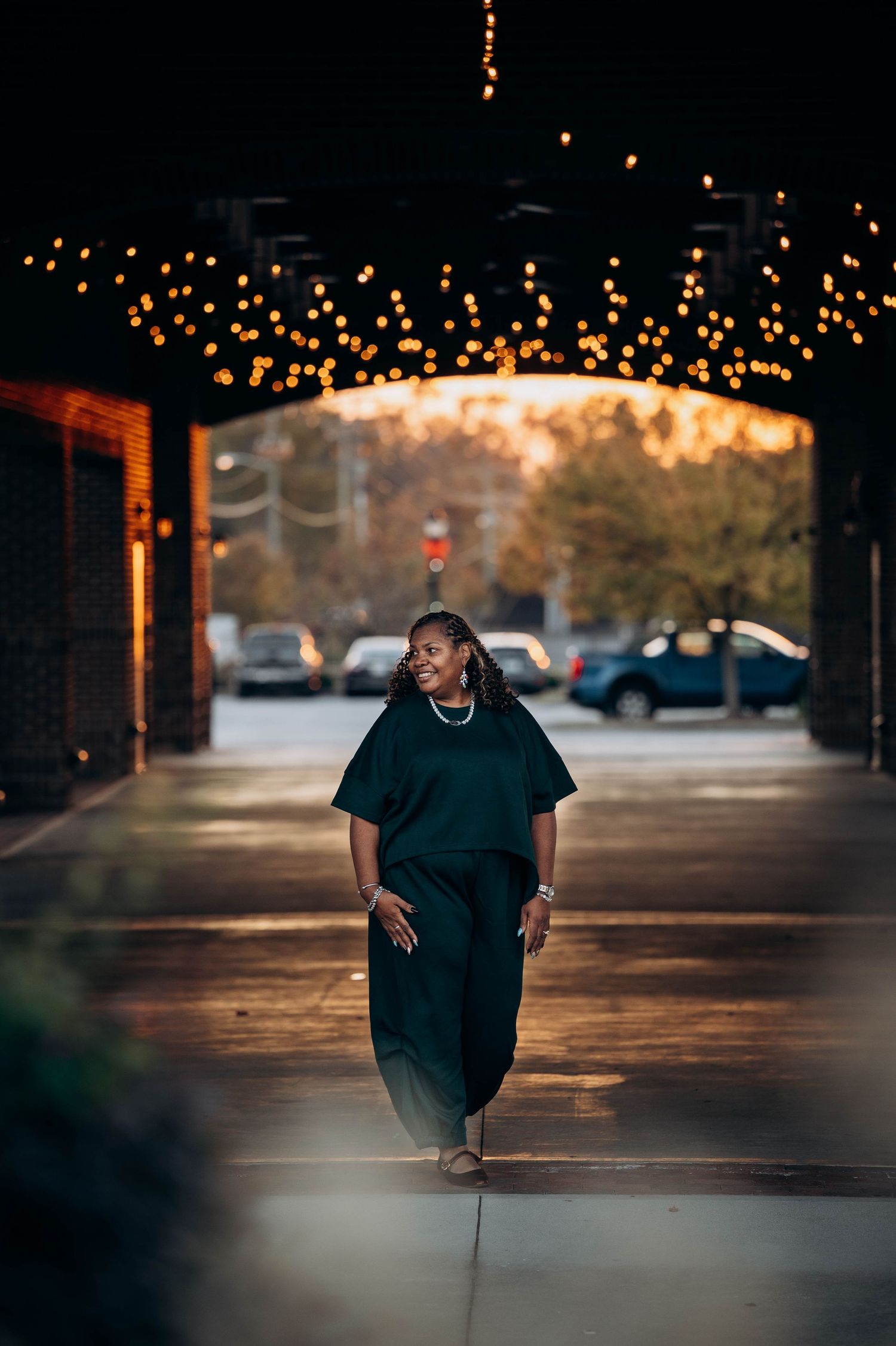 A person in dark clothing walks under string lights in an urban walkway at dusk.