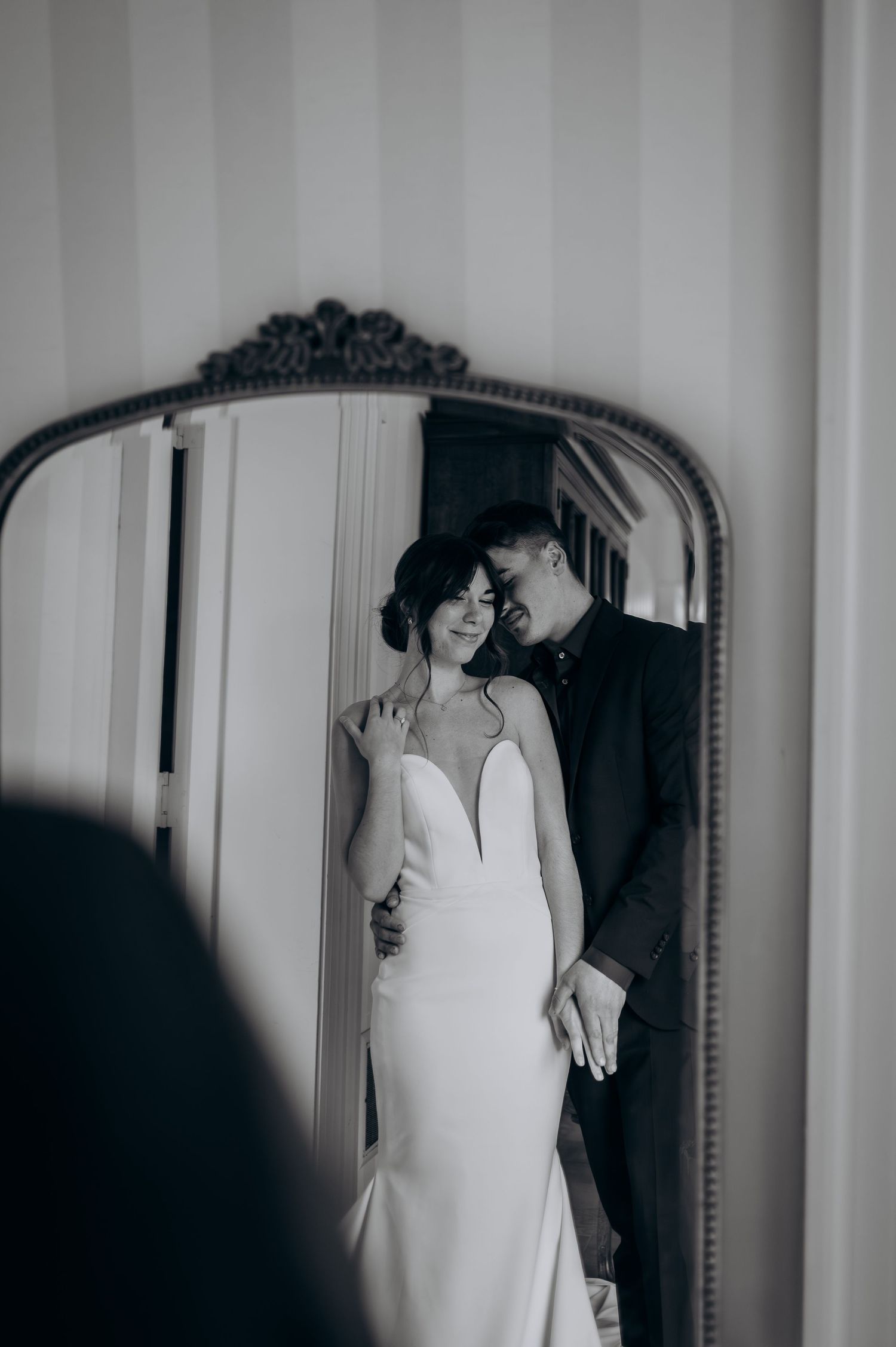 Black and white wedding photo of bride and groom reflected in ornate vintage mirror, bride in elegant gown, groom in dark suit.