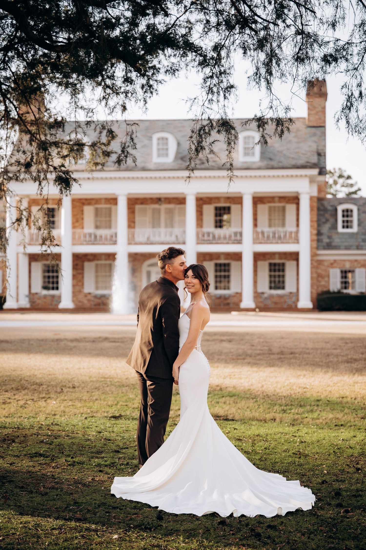 Bride and groom share a kiss on the lawn of a grand brick colonial mansion at golden hour.