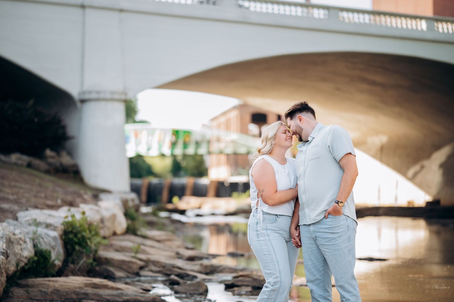 Couple sharing a romantic moment under a stone bridge by a river, dressed in casual white and light blue outfits.