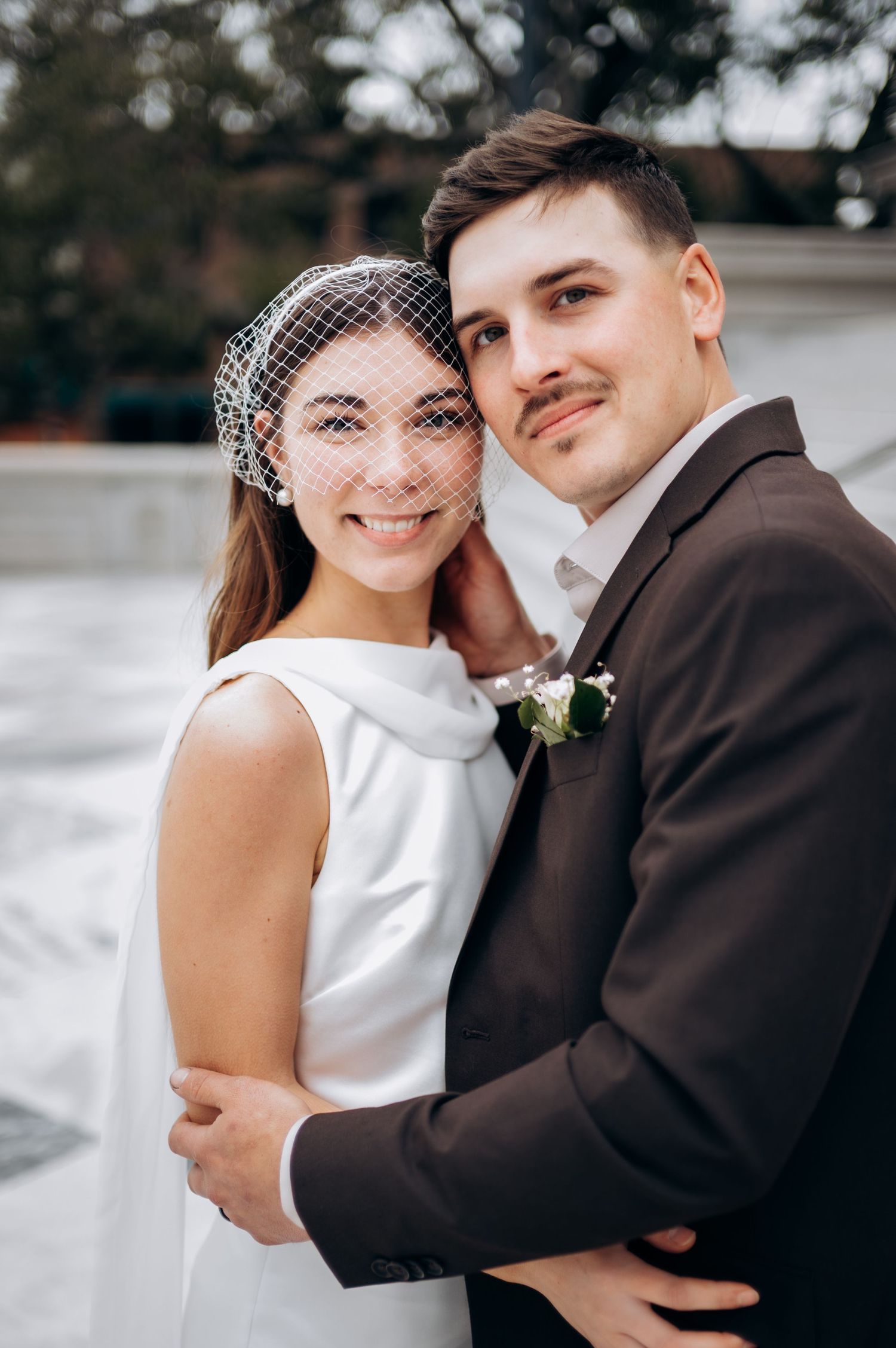 A couple in wedding attire embraces outdoors with a soft focus background of trees and foliage.