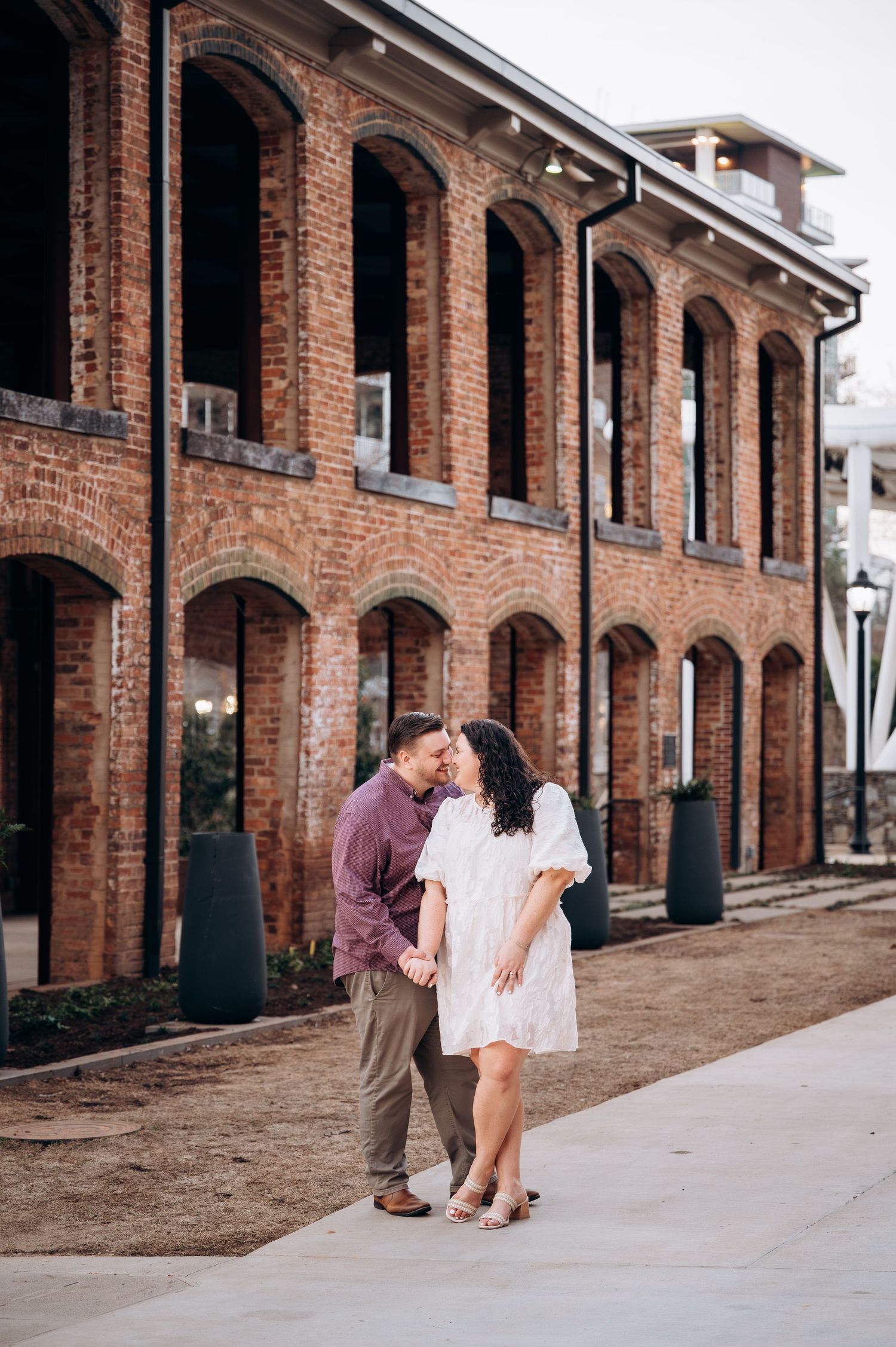 A couple in romantic pose stands together against a backdrop of rustic brick arches and columns.