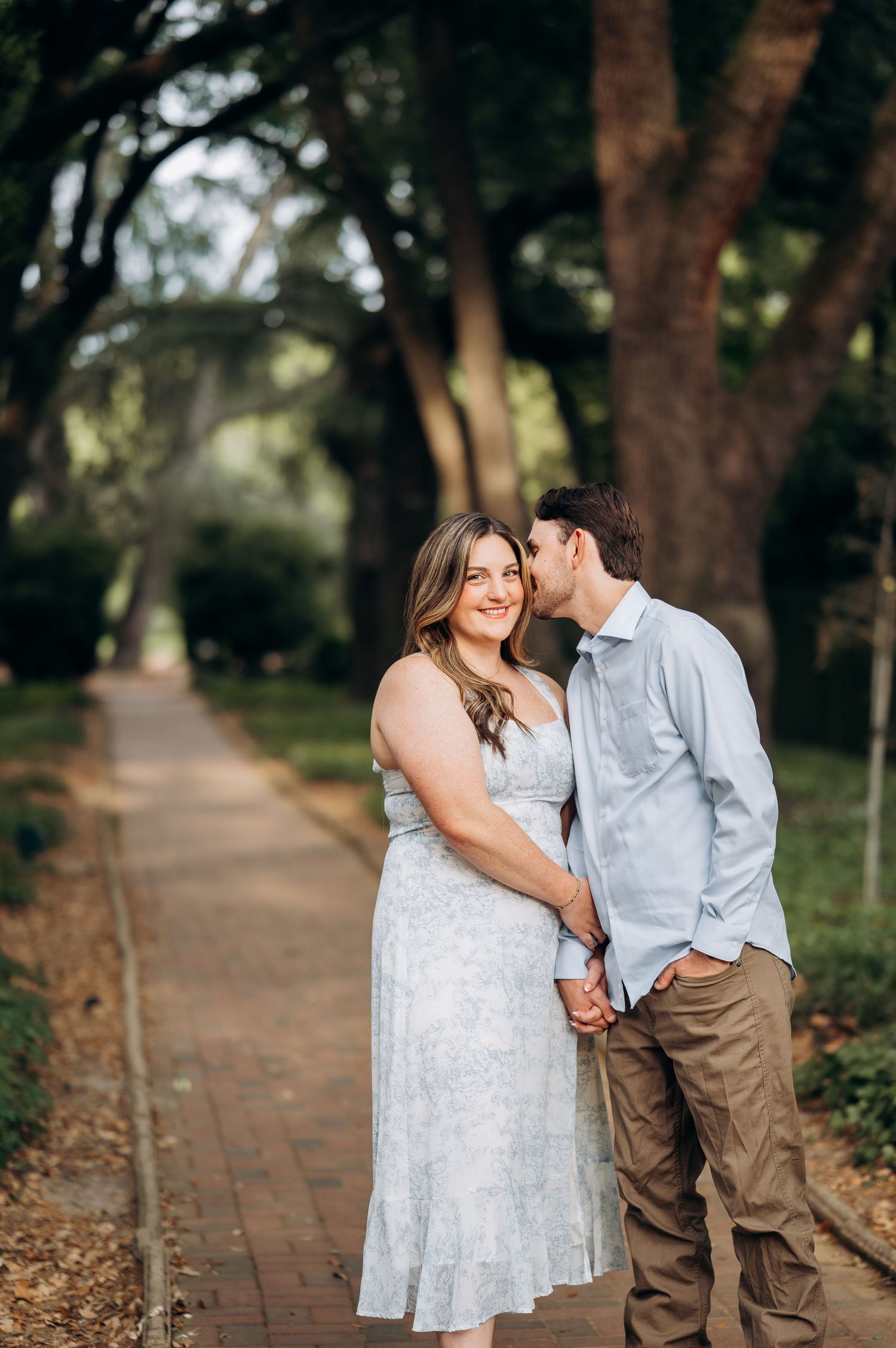 Couple holding hands on tree-lined brick path, man kissing womans cheek during engagement photo session.