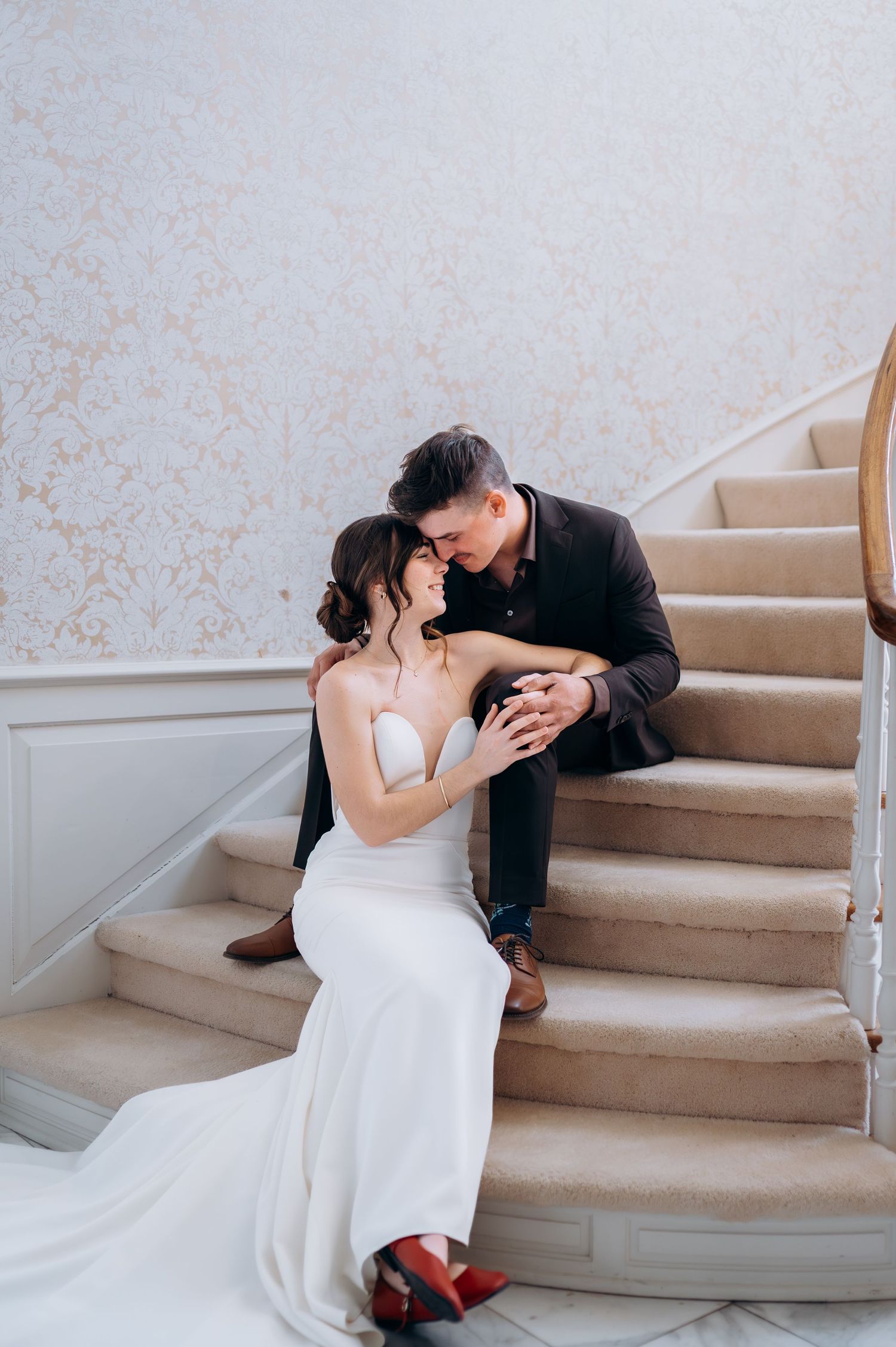 Bride and groom sit closely on elegant carpeted staircase, sharing a romantic moment in a grand venue.