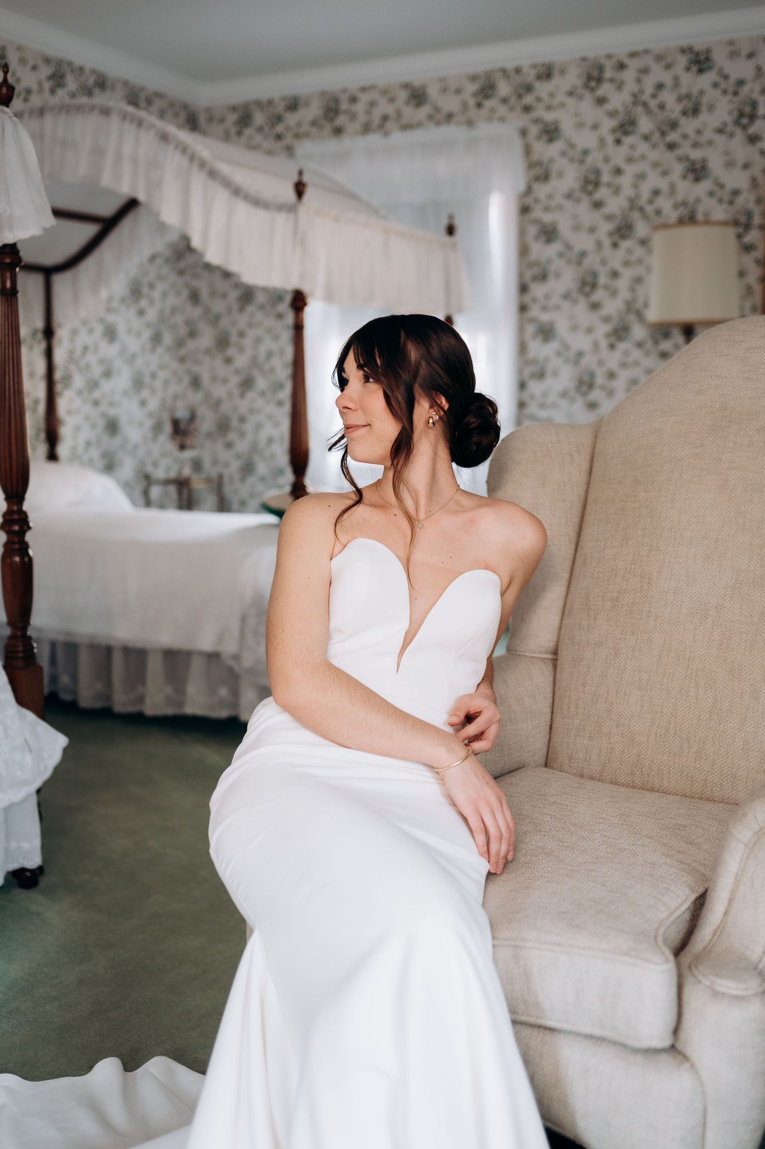 Bride in strapless white mermaid gown sits gracefully in a vintage armchair in an elegant floral bedroom.