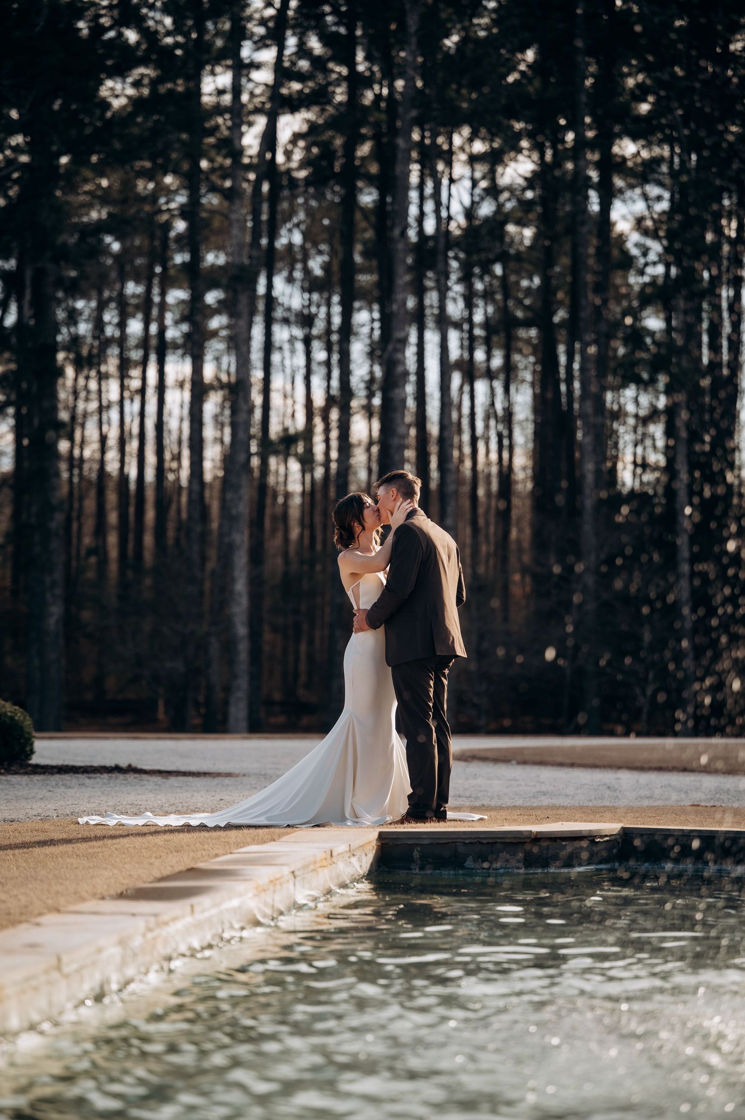 Bride and groom kissing beside a reflective pool with tall pine trees behind them.