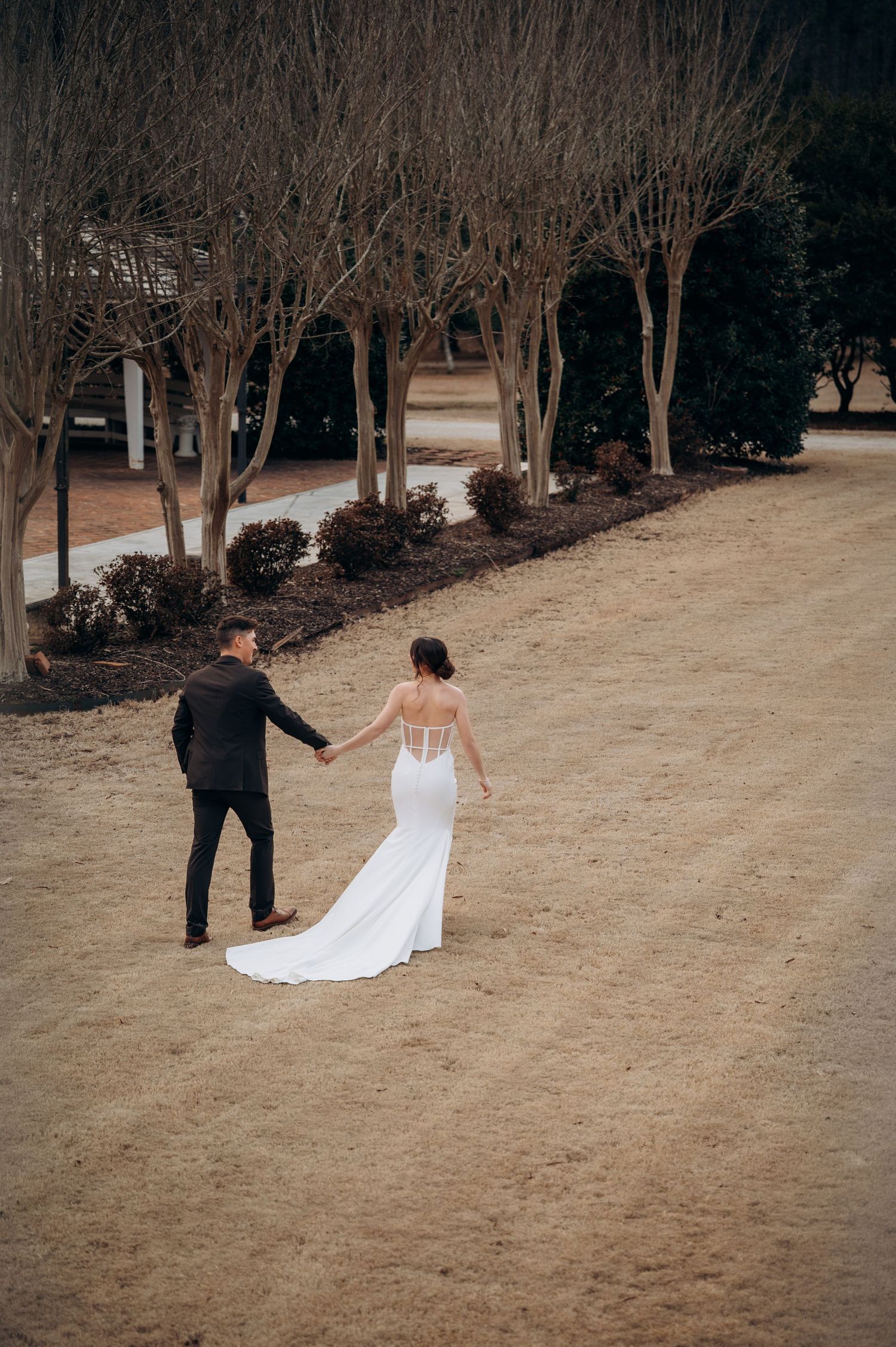 Bride and groom walk hand-in-hand along a sandy path lined with bare winter trees.
