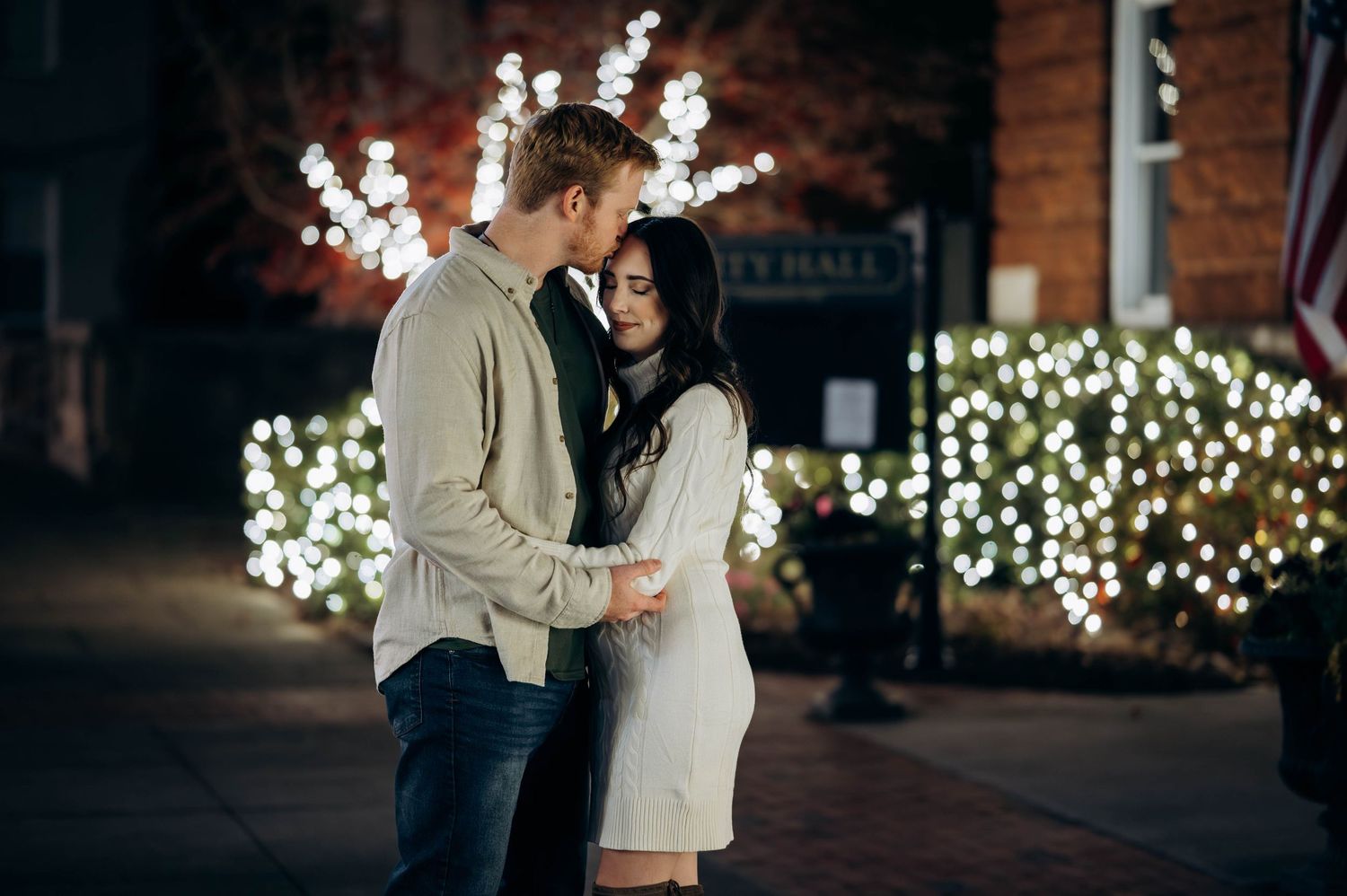 Romantic couple holding hands near festive Christmas lights outside City Hall at night.