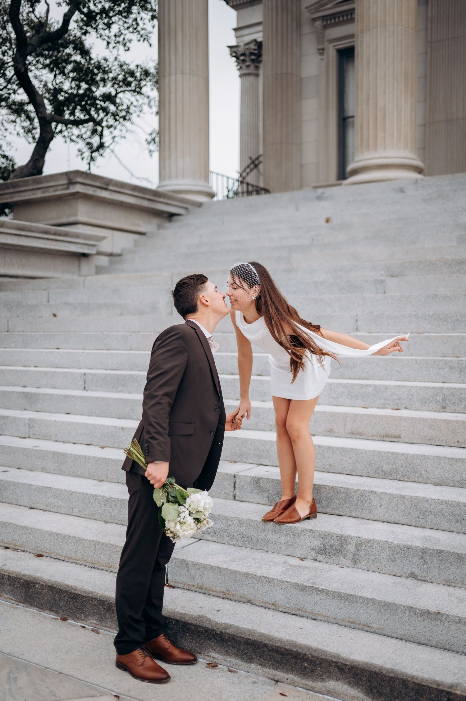 Couple sharing a romantic kiss on grand stone steps, groom holding white bouquet.