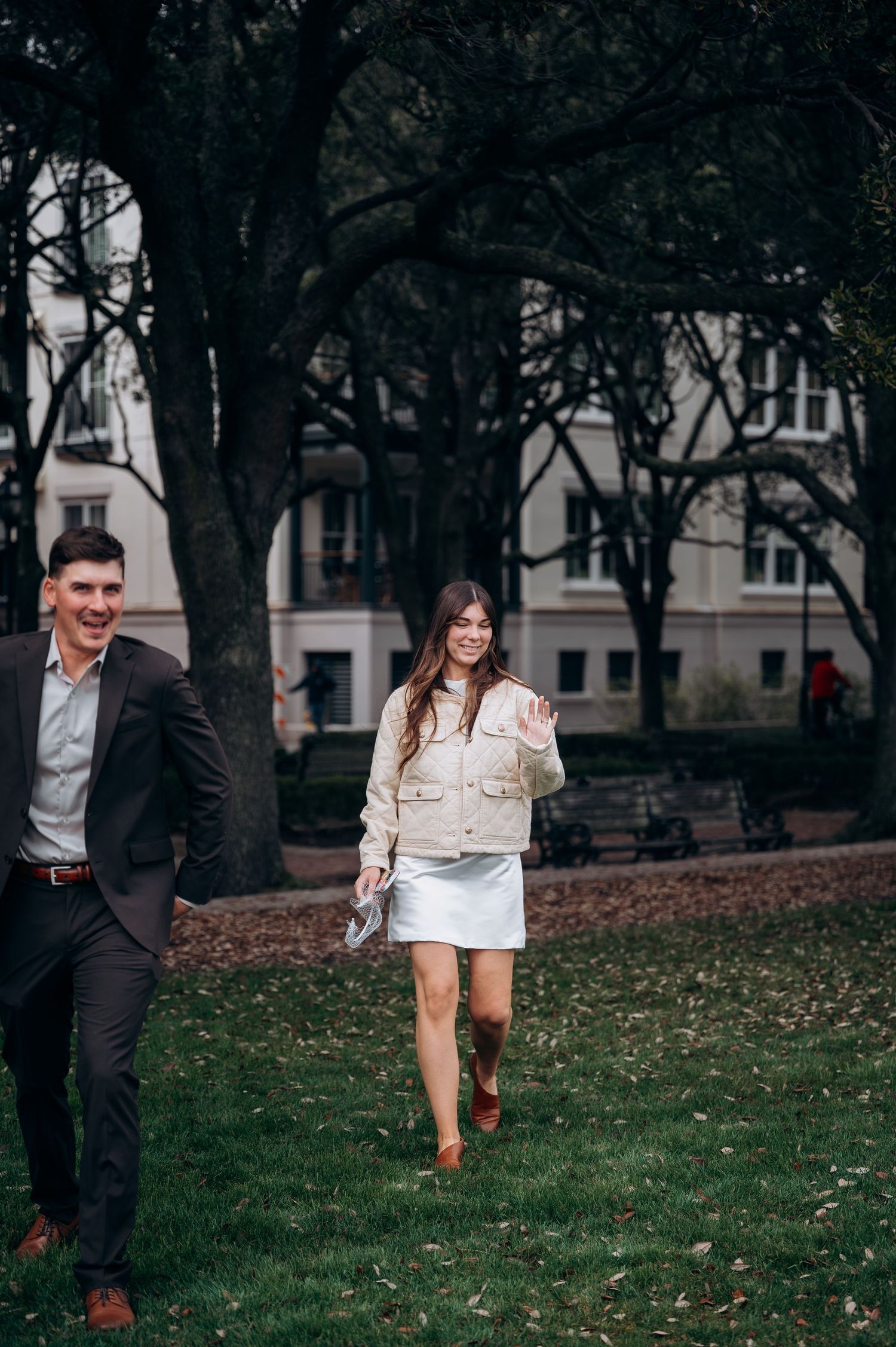 Happy couple walking through green park, woman in cream jacket and white skirt, man in dark suit.