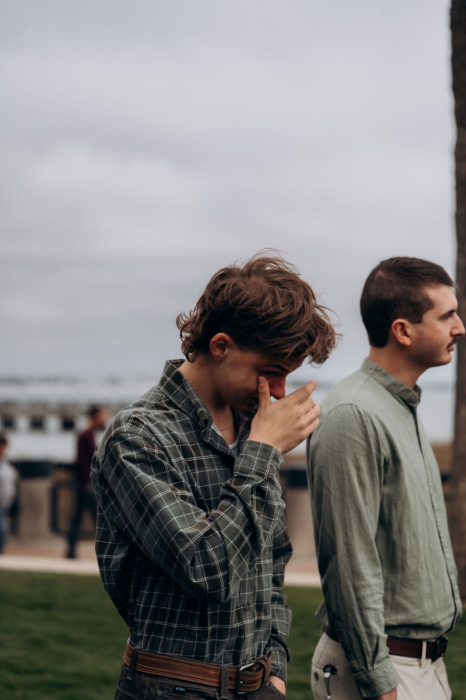 Two men standing outdoors near waterfront, one in plaid shirt looking down, other in sage green shirt.