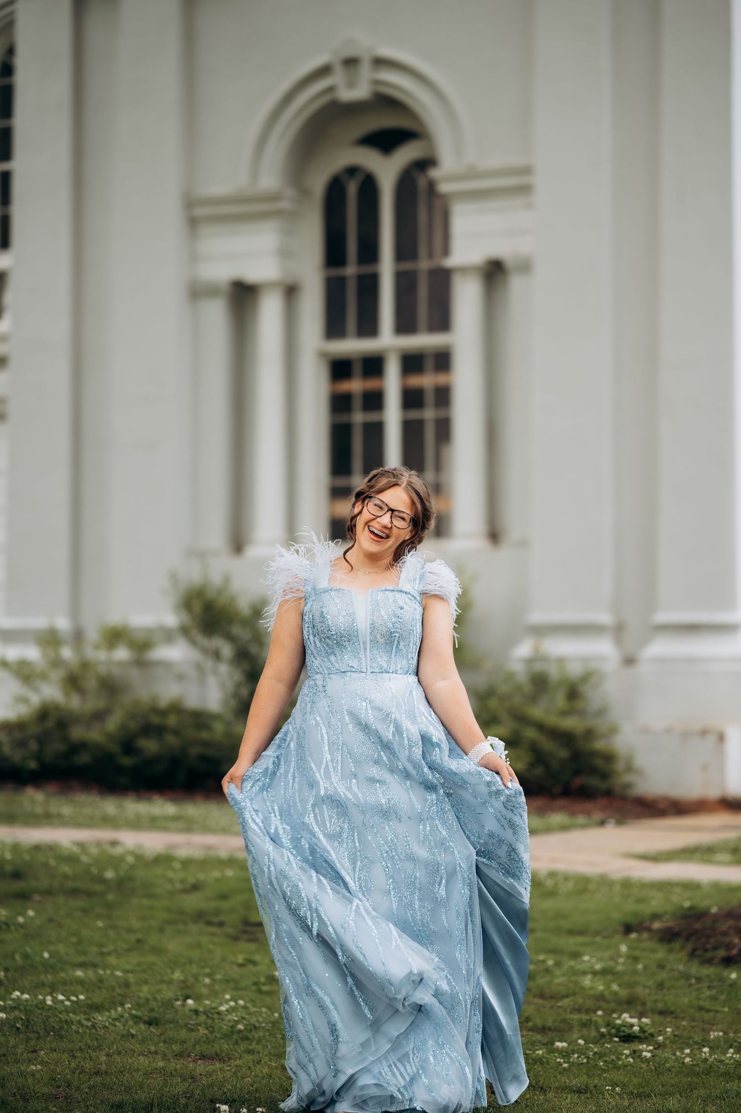 Young woman in flowing light blue tulle gown laughs joyfully outside white classical building.