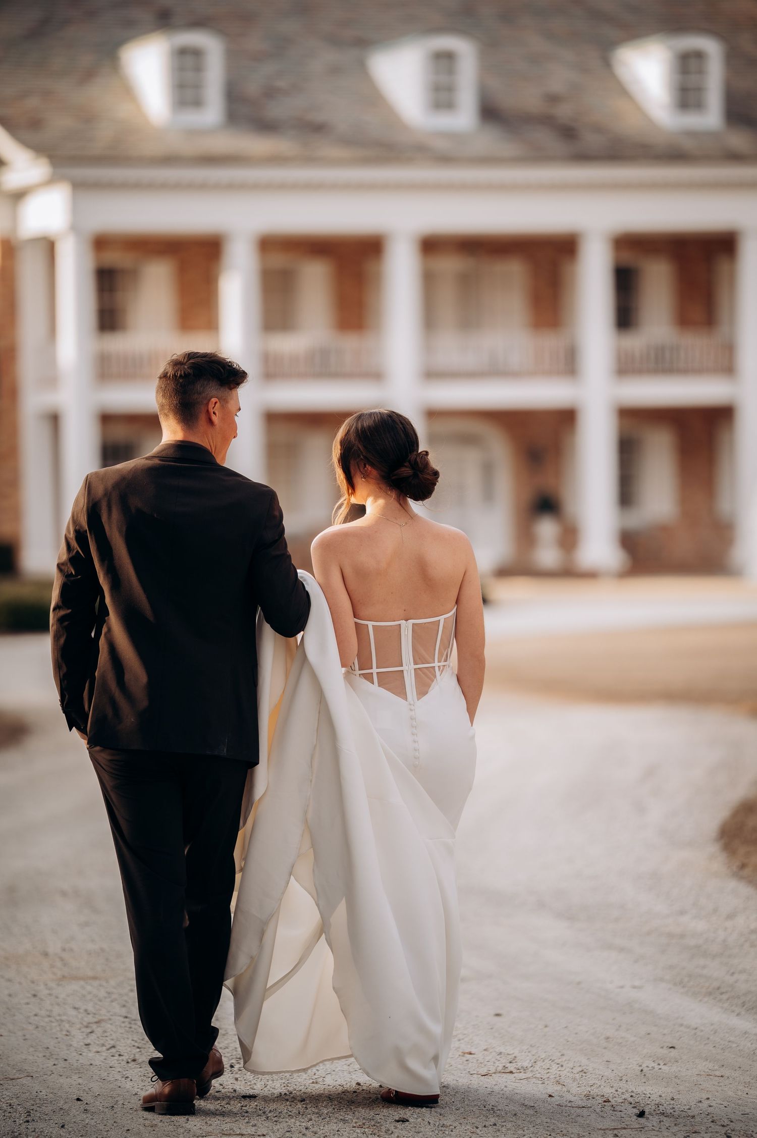 Elegant wedding couple walking along a colonial style building with white columns and gravel path.