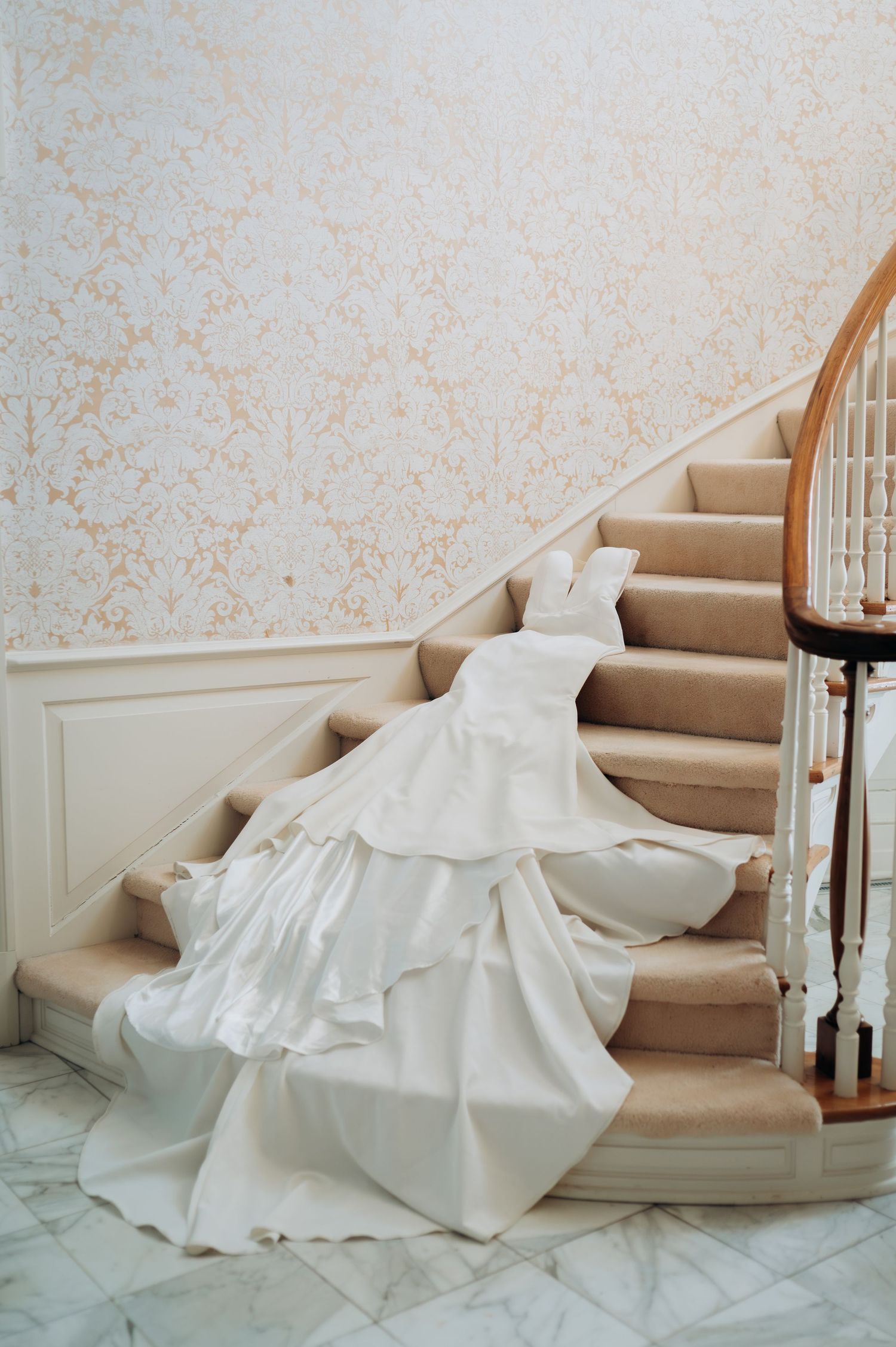 A flowing white wedding dress elegantly draped across beige carpeted stairs with floral wallpaper in the background.