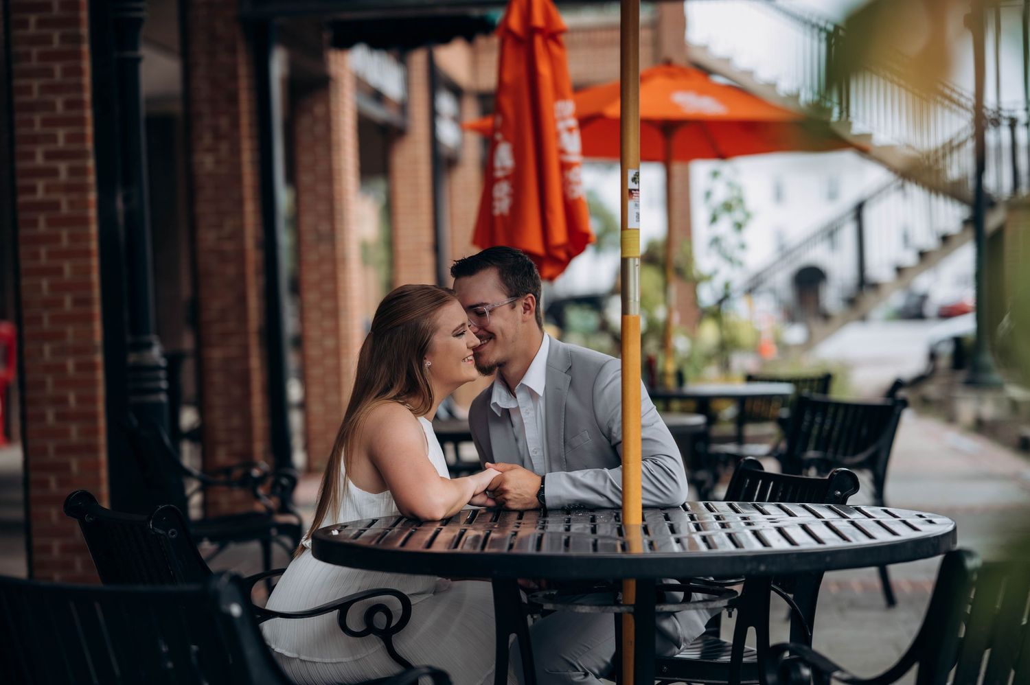 Romantic couple shares tender moments on a brick-lined sidewalk with orange umbrellas in background.