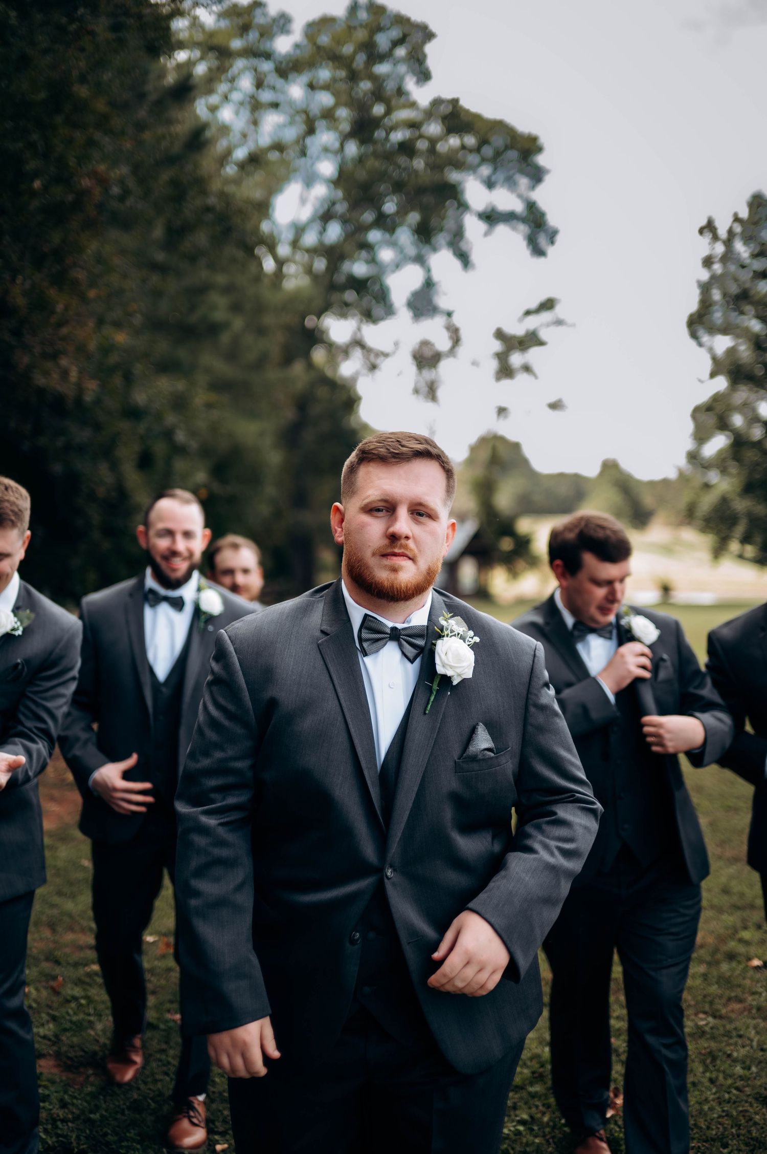 Wedding party in black tuxedos and bow ties poses on grassy lawn.