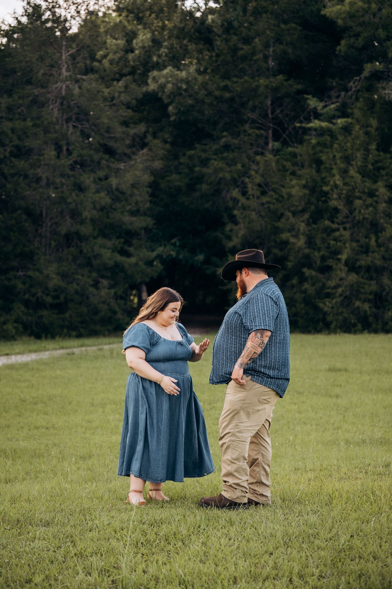 Couple standing in green field, woman in blue dress touching her belly while man in cowboy hat watches.