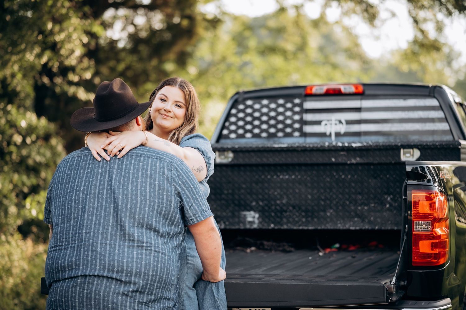 Woman hugging a man in a cowboy hat beside a black pickup truck with an American flag rear window.