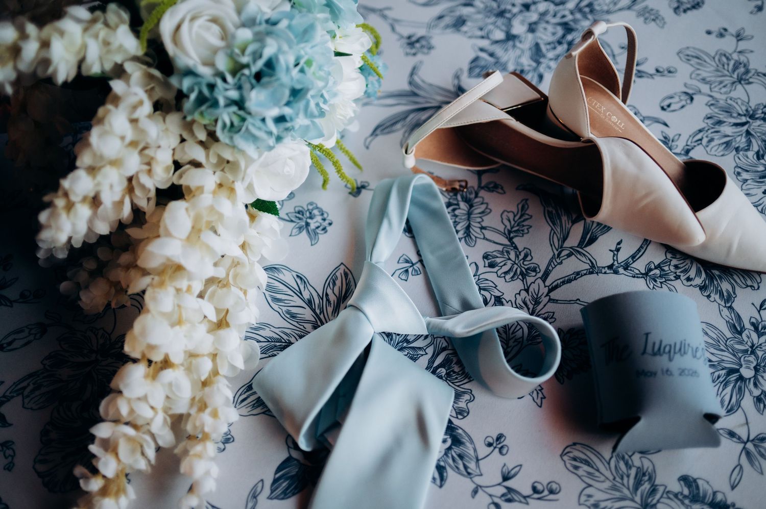 Wedding flat lay with blush heels, blue ribbon, and white floral bouquet.