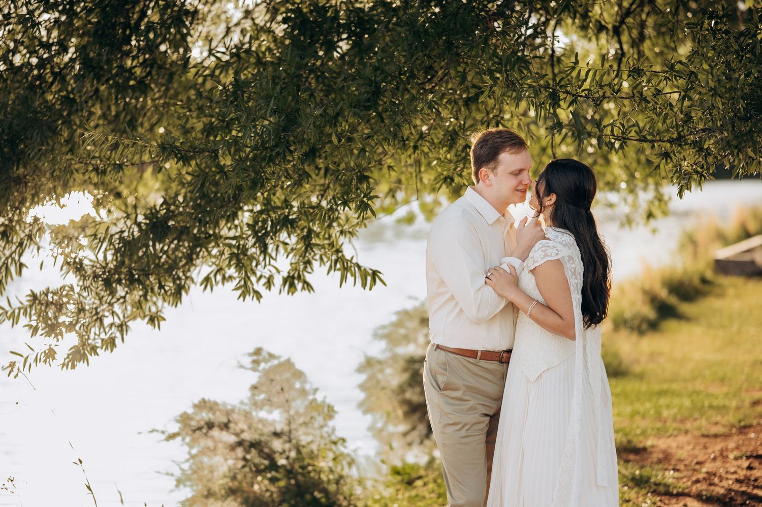Couple sharing a romantic moment under weeping willow tree by waterfront, bride in white dress and groom in tan suit.
