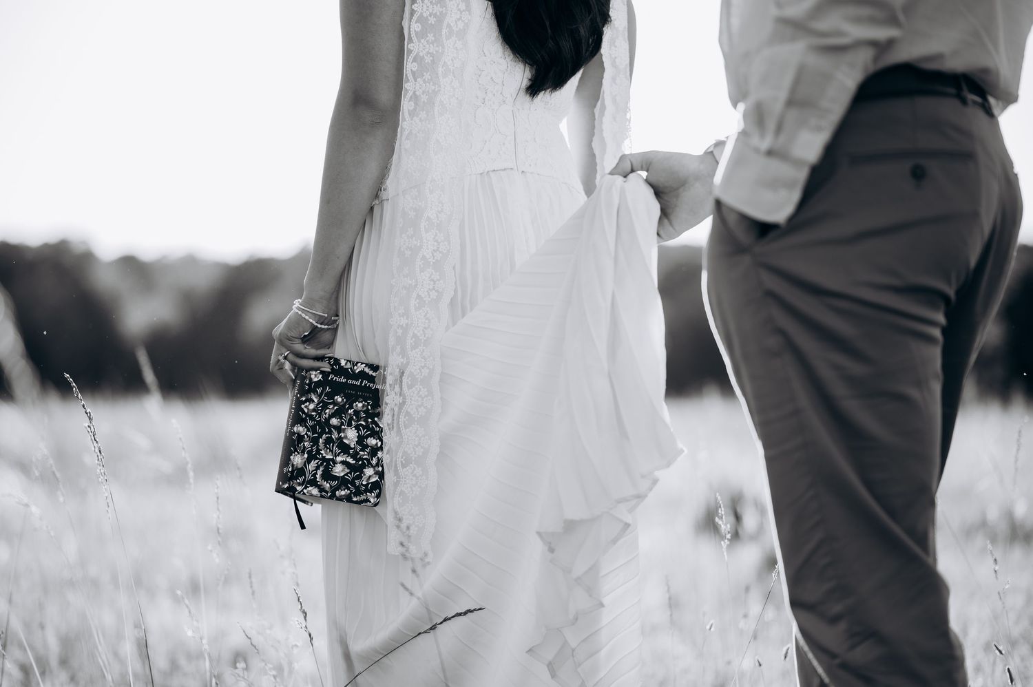 Black and white photo of a bride in white dress holding a clutch, walking hand in hand with groom in a field.