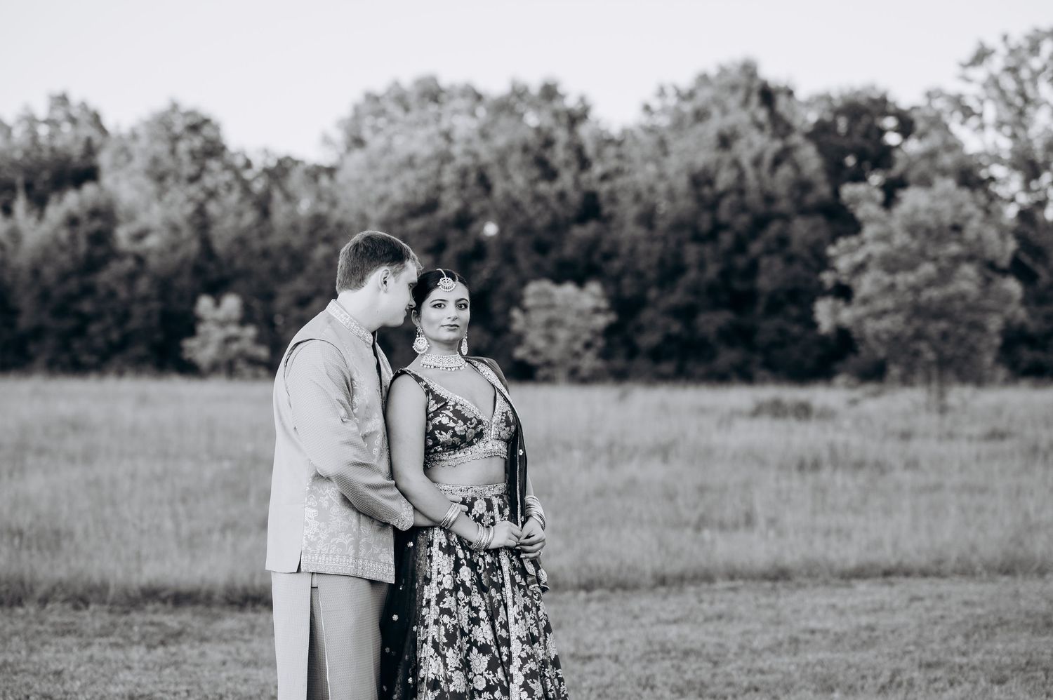 Black and white photo of a couple standing in a field, sharing a tender moment with trees in the background.