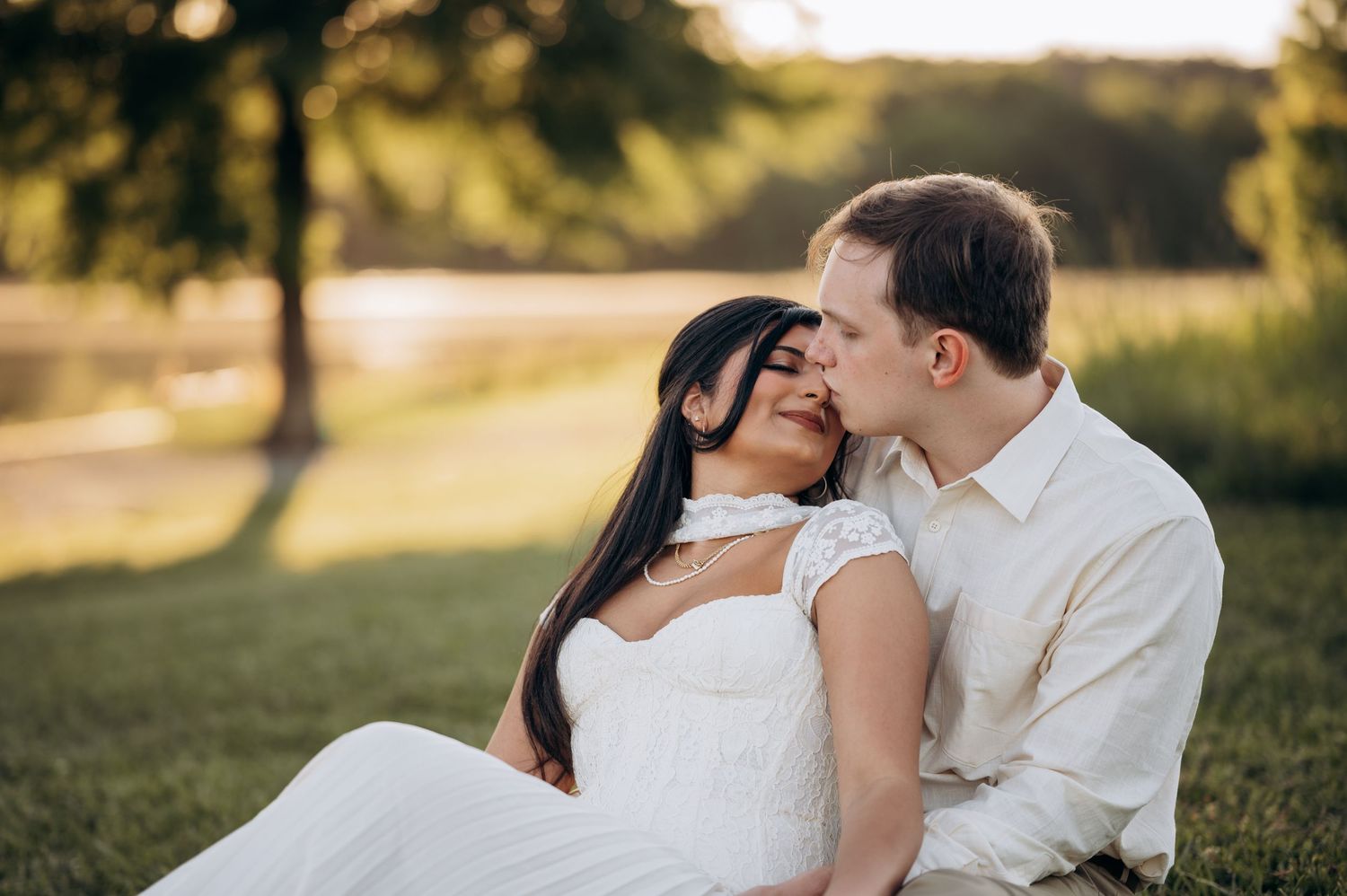 Romantic couple in white attire sitting on grass at sunset, sharing a tender moment near a serene lake.