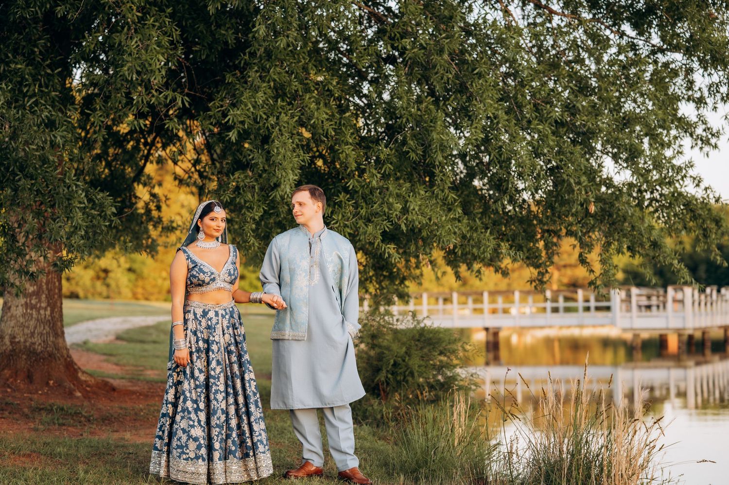 Couple in traditional Indian attire standing by a serene lake surrounded by lush green trees during golden hour.