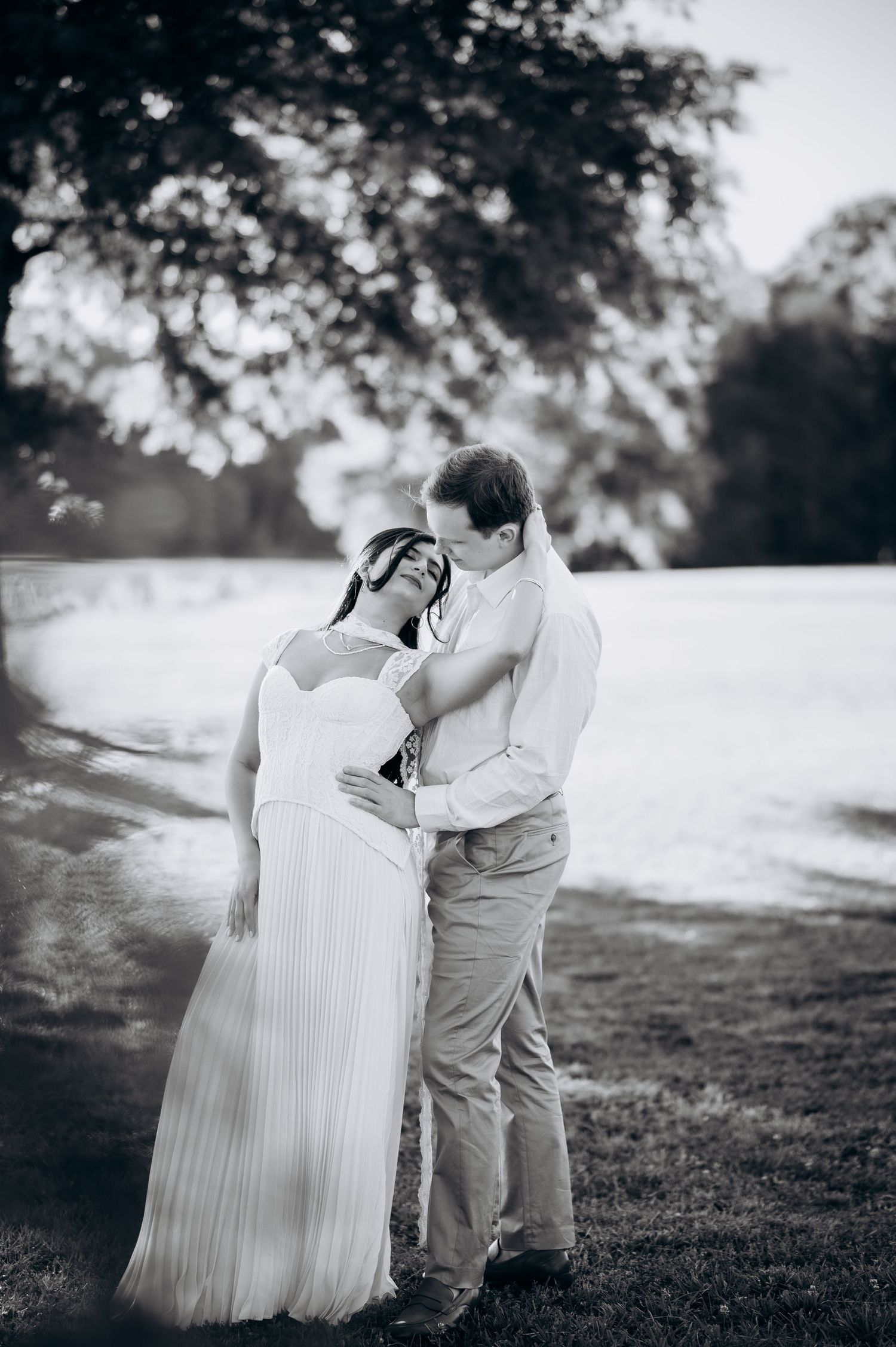 Black and white photo of a couple embracing outdoors, woman in white gown, man in dress shirt, trees in background.