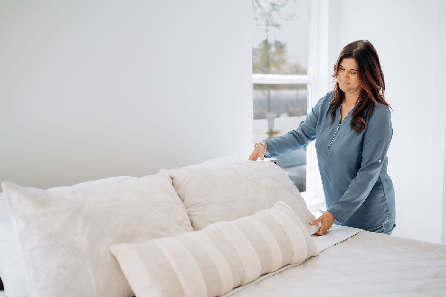 Woman in blue pajamas making bed with white pillows and striped cushion in bright minimalist bedroom.
