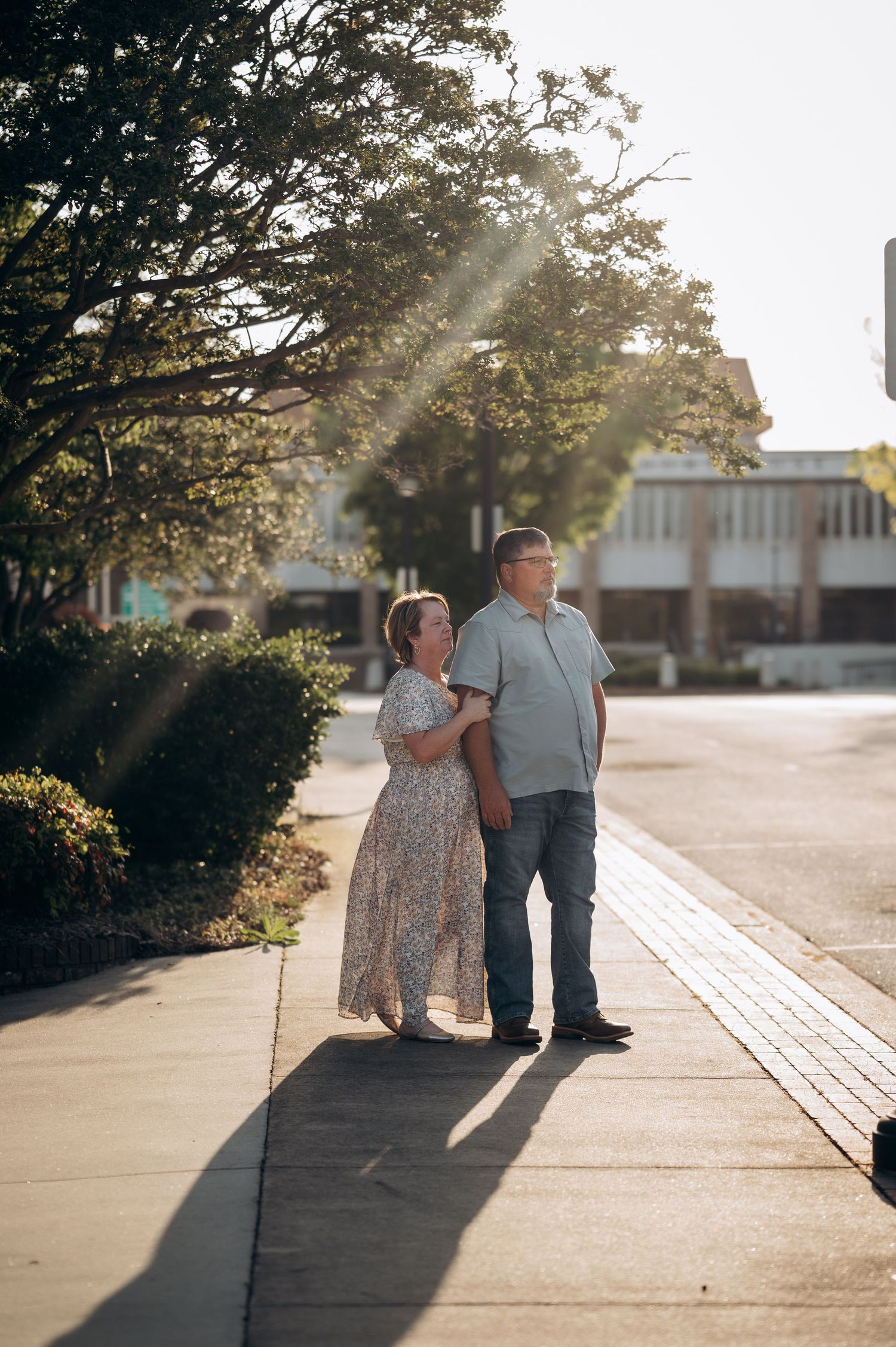 Couple standing together on a sunlit sidewalk, surrounded by trees and buildings, bathed in warm golden sunlight.