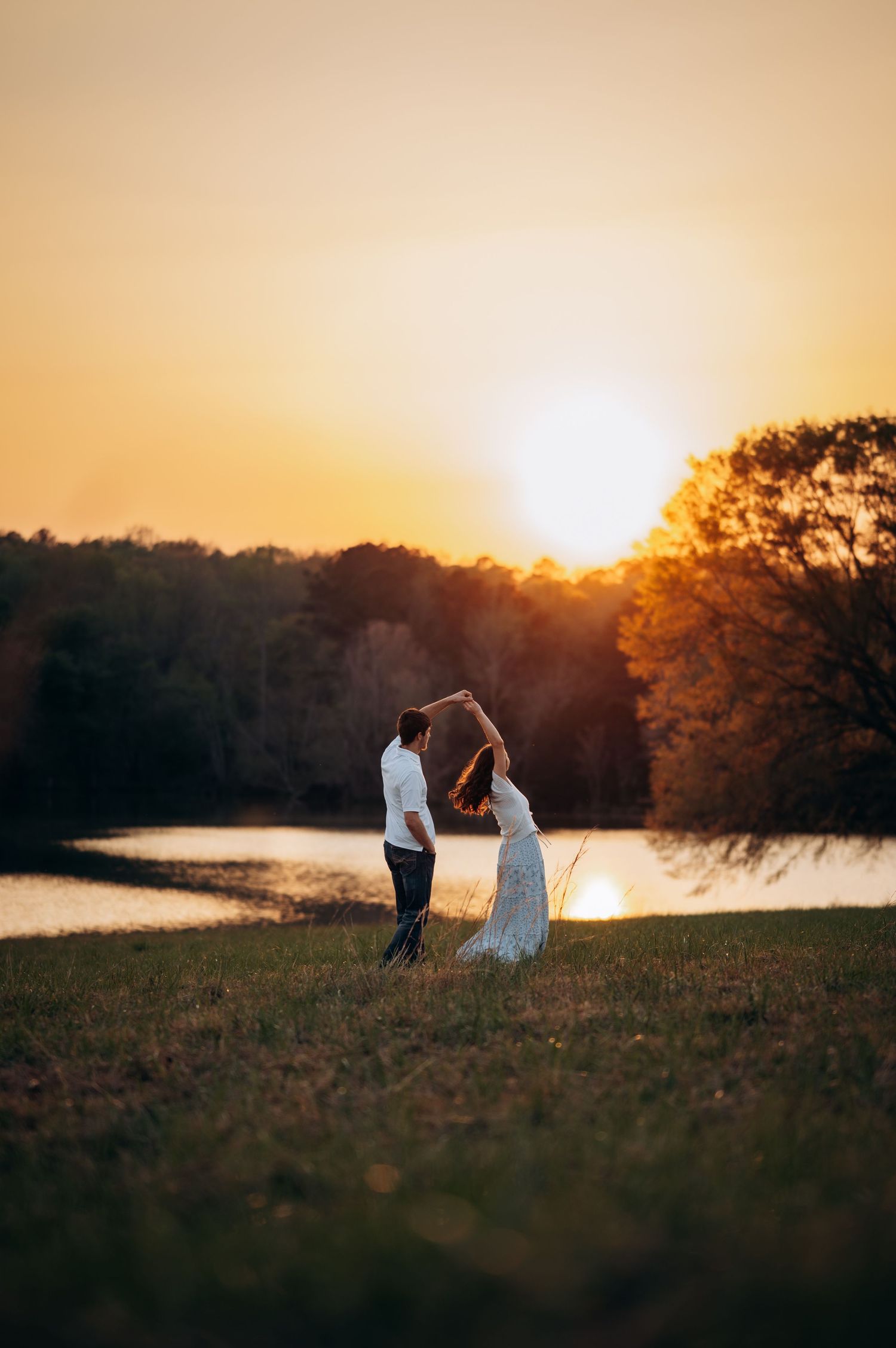 Couple dancing at sunset by a lake, bride in white gown, golden hour light, romantic outdoor wedding photo.