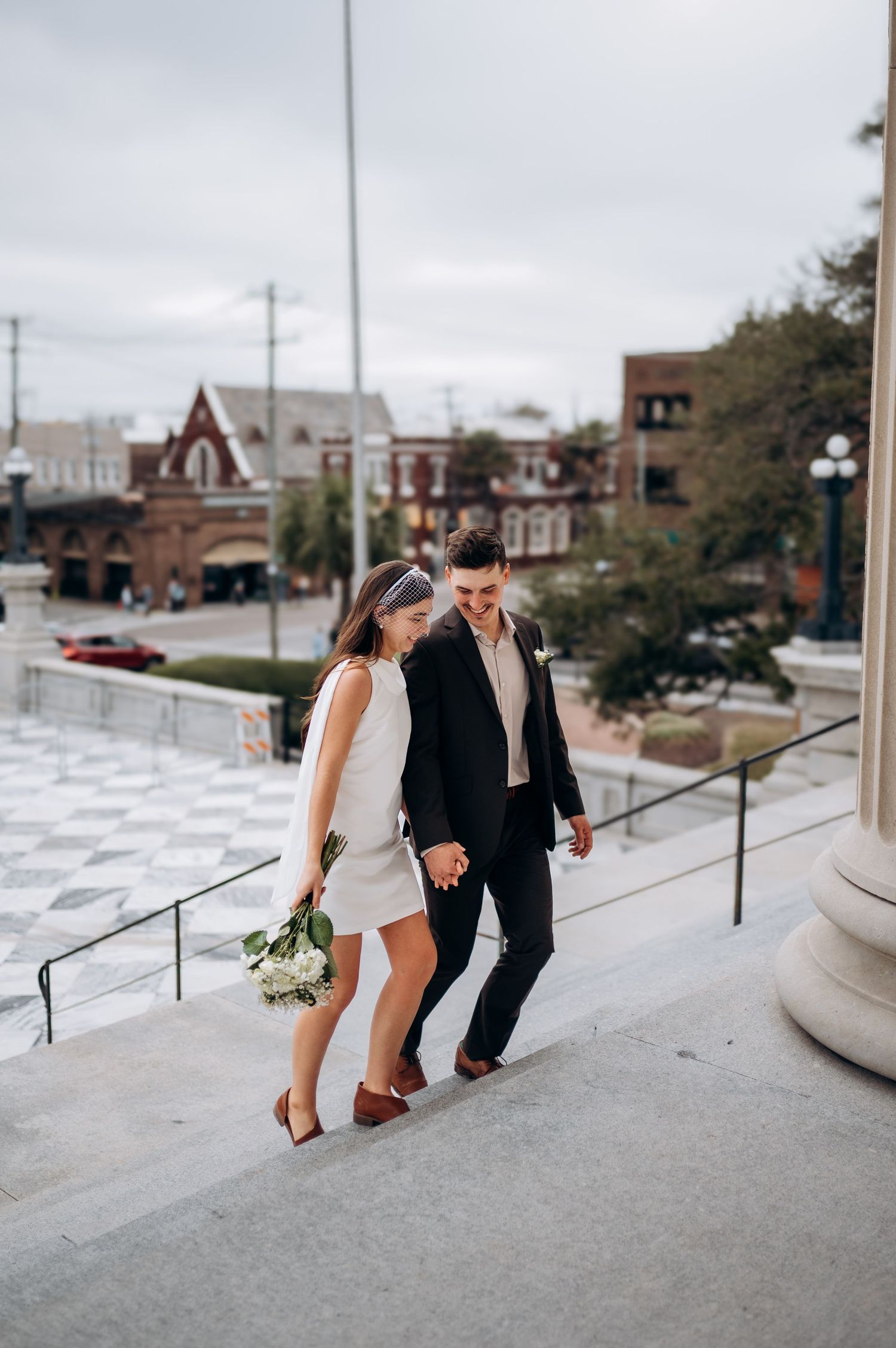 Couple in wedding attire holding hands walking down outdoor steps with bouquet, urban buildings in background.