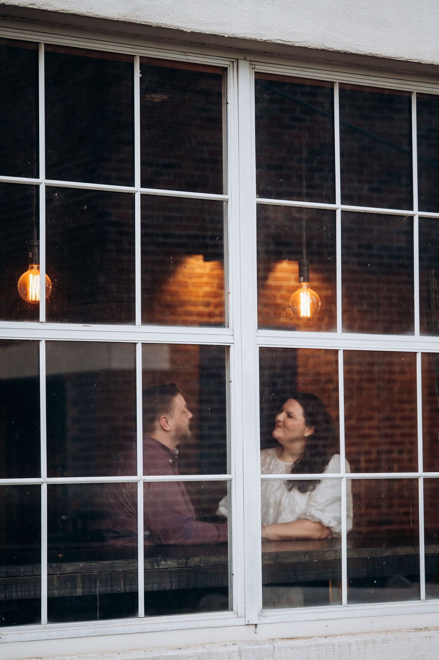 Two people sit at a table by large windows in a dimly lit restaurant with hanging pendant lights and exposed brick walls.